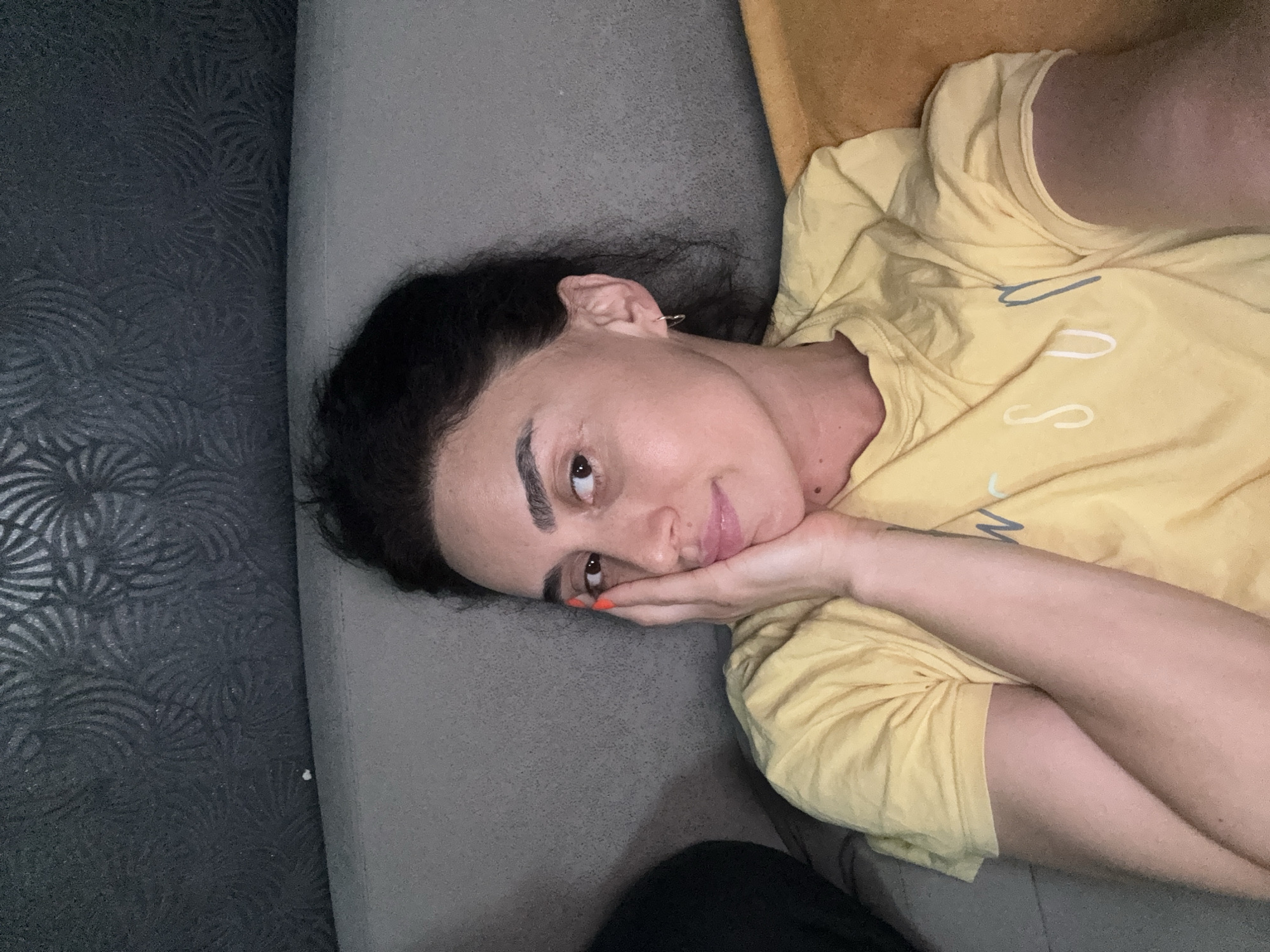The image shows a woman with dark hair, wearing a yellow t-shirt, resting on a gray sofa. She is holding her right hand to her face, with her fingers on her cheek. A dark, textured wallpaper is visible in the background, and an orange pillow is on the right side of the frame. The location is the city of Tbilisi, Georgia.