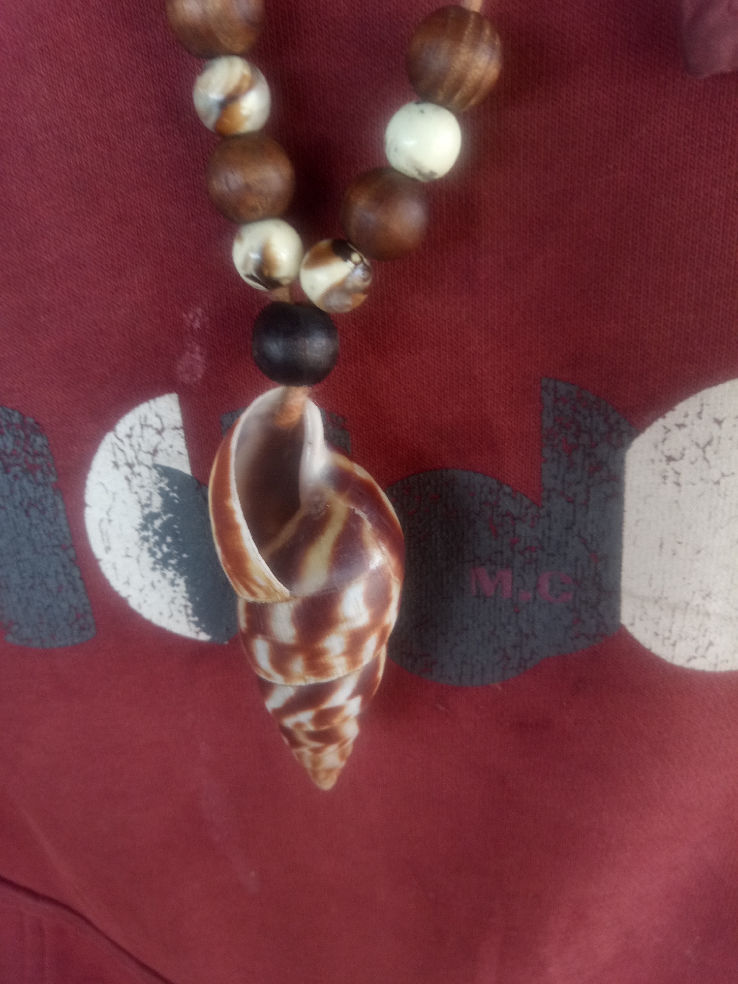 A close-up, slightly angled shot showcases a necklace made of various beads, including wooden spheres in shades of brown, white, and a dark, possibly black, bead, all strung on a thin cord. The beads are arranged in a curved line. Suspended from the dark bead is a large, spiral seashell pendant, exhibiting brown and white stripes. The necklace rests against a deep red fabric, likely a garment, which is subtly patterned with abstract, grey, and white shapes. The lighting is even, and the focus is sharp on the necklace and the immediate background fabric. The image does not depict any specific urban environment or geographical location.
