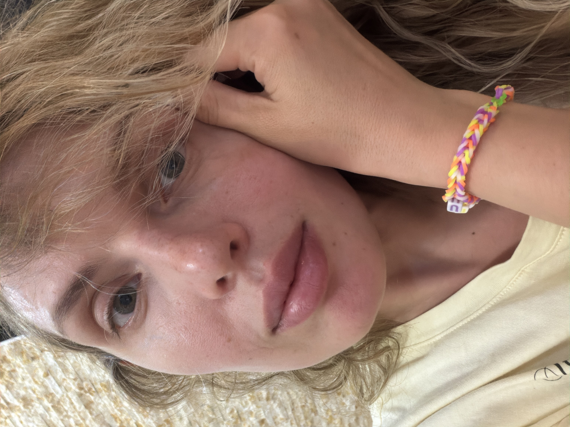 A close-up, slightly rotated portrait of a fair-skinned individual with blonde hair, focusing primarily on their face and the upper portion of their body. The individual's left hand is positioned near their face, with a colorful braided bracelet visible on their wrist. They are wearing a light yellow top. The background is indistinct but appears to be a textured surface. There is no indication in the image as to its location in the city of Budva, Montenegro.