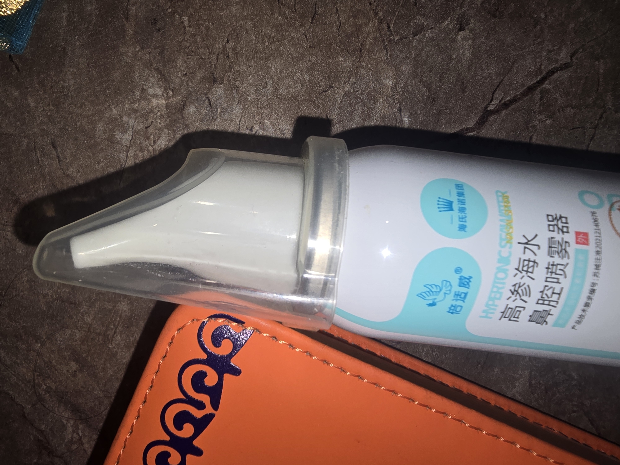 The image is an overhead shot of a white spray bottle with a clear plastic nozzle, resting on a dark, textured surface. The bottle has Chinese text and an illustration of a bird. To the right of the bottle is a bright orange, textured object with an ornate dark blue design along its edge, which appears to be a case or cover. The scene is lit, casting shadows. There is no indication of the location in Ulan-Ude, Russia.