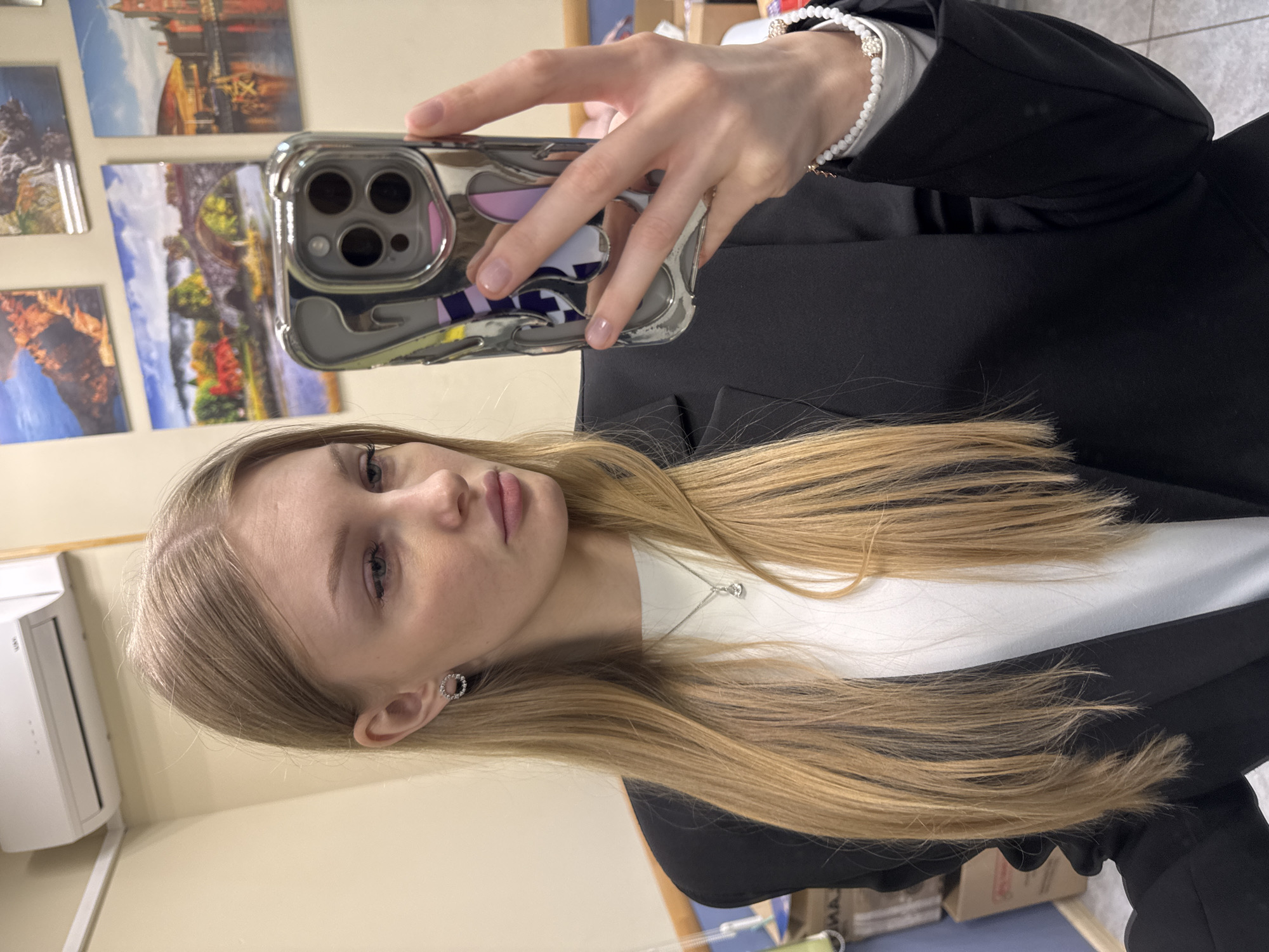 A young woman with long, straight blonde hair and light-colored eyes is captured in a selfie. She wears a black blazer over a white top, accessorized with a silver chain necklace featuring a single clear pendant, and small circular earrings with clear stones. Her left hand holds a smartphone, clad in a reflective silver/chrome case with a textured, abstract design and a prominent triple camera lens setup. Her right wrist sports a white beaded bracelet with a decorative clasp.
The woman holds the phone up, seemingly using its front camera for the selfie, with her gaze directed at the screen. Her expression is neutral to slightly contemplative.
The background is a light, cream-colored wall adorned with several framed or mounted landscape and cityscape photographs. These pictures depict scenes like a city with a large bridge, a vibrant autumn landscape with a bridge, and possibly other urban or natural vistas. To the left, a white wall-mounted air conditioning unit is visible. Partial text "DEA" and potentially "M" from "MIDEA" are discernible on the unit, along with other unreadable smaller text. The overall setting appears to be an indoor space, possibly an office or public building in Perm, Russia, given the provided location context. The lighting suggests daytime.