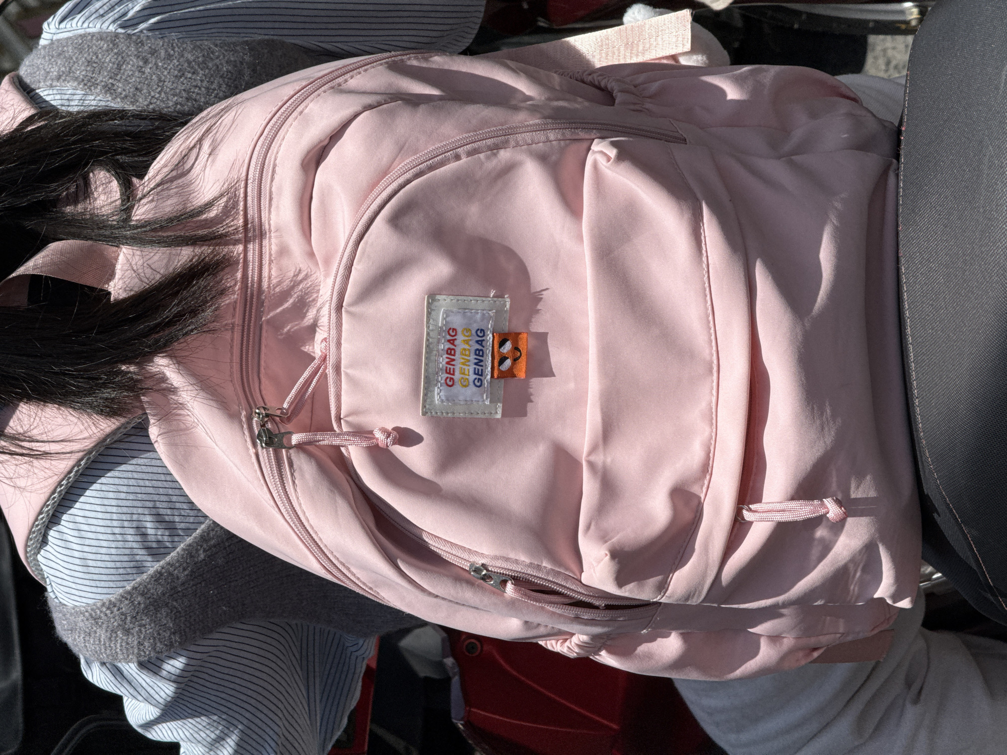 A pink backpack is prominently featured in the image. The backpack has a tag labeled "GENBAG" in multiple colors. The bag is resting on a surface with a dark gray seat or cushion visible on the right side. To the left, a person is wearing a striped shirt and a gray sweater. Long, dark hair is visible on the left side of the backpack. The setting or city is not clear from the image.