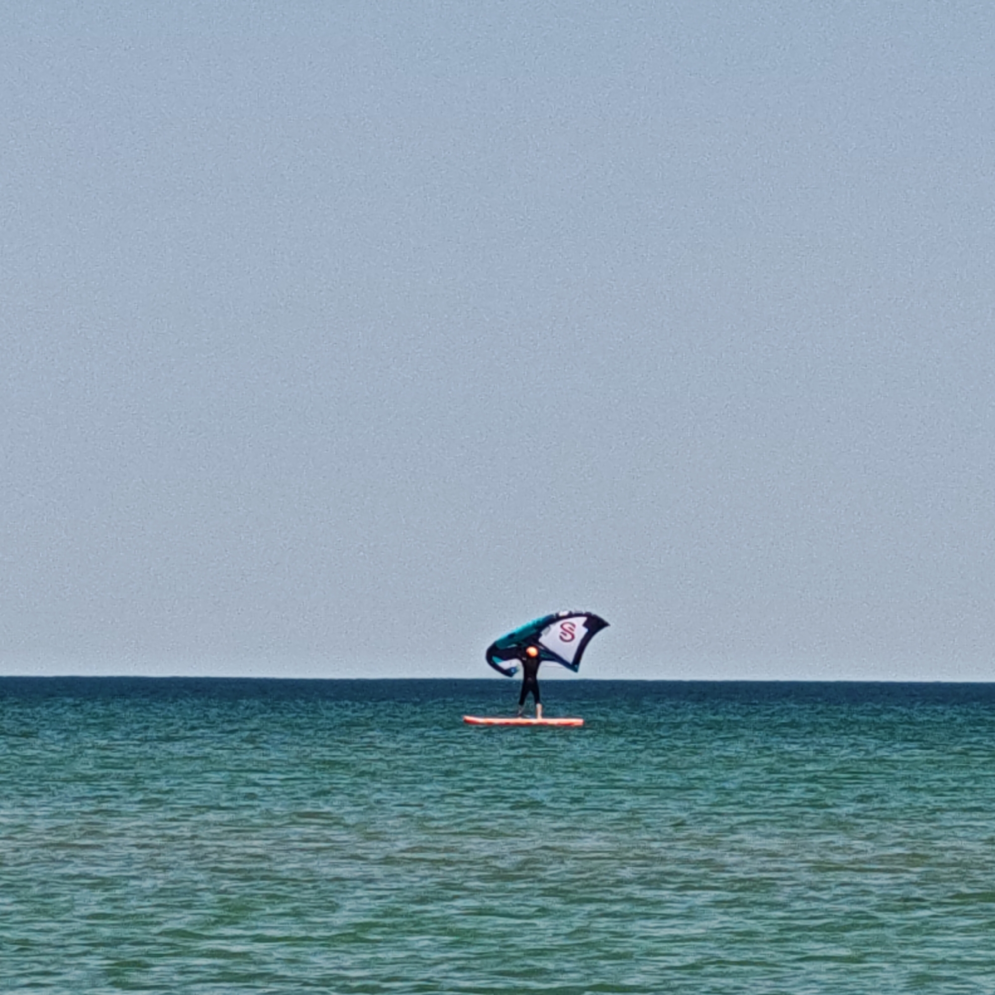 A distant shot shows a person on a paddleboard on the sea near городское поселение Щелкино, Ukraine. The person is holding a windsurfing sail. The sea is aqua green, and the sky is a pale grey-blue. The horizon is level.