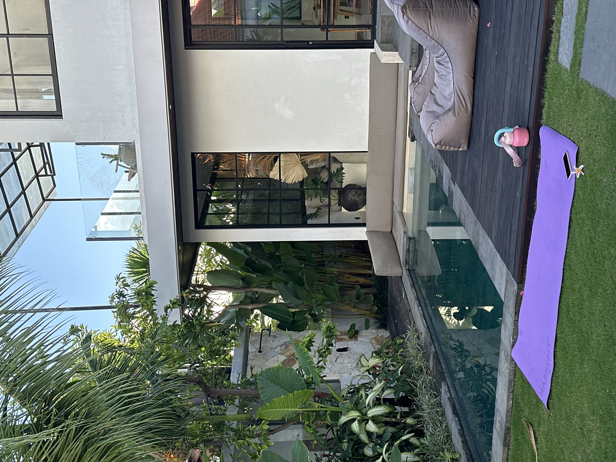The photograph depicts a scene in North Kuta, Indonesia, featuring an outdoor patio area. The patio includes dark wood decking and a light-colored stone or concrete patio section. A lounge chair and a light-colored sofa are positioned on the patio. A narrow pool or water feature runs alongside the patio area, reflecting the surrounding vegetation. Tropical foliage is abundant, including various plants and trees. A purple yoga mat, a water bottle, and a phone are positioned on the grass adjacent to the patio. The architecture includes modern elements with large windows and a glass roof section. The overall scene suggests a tranquil, tropical setting.