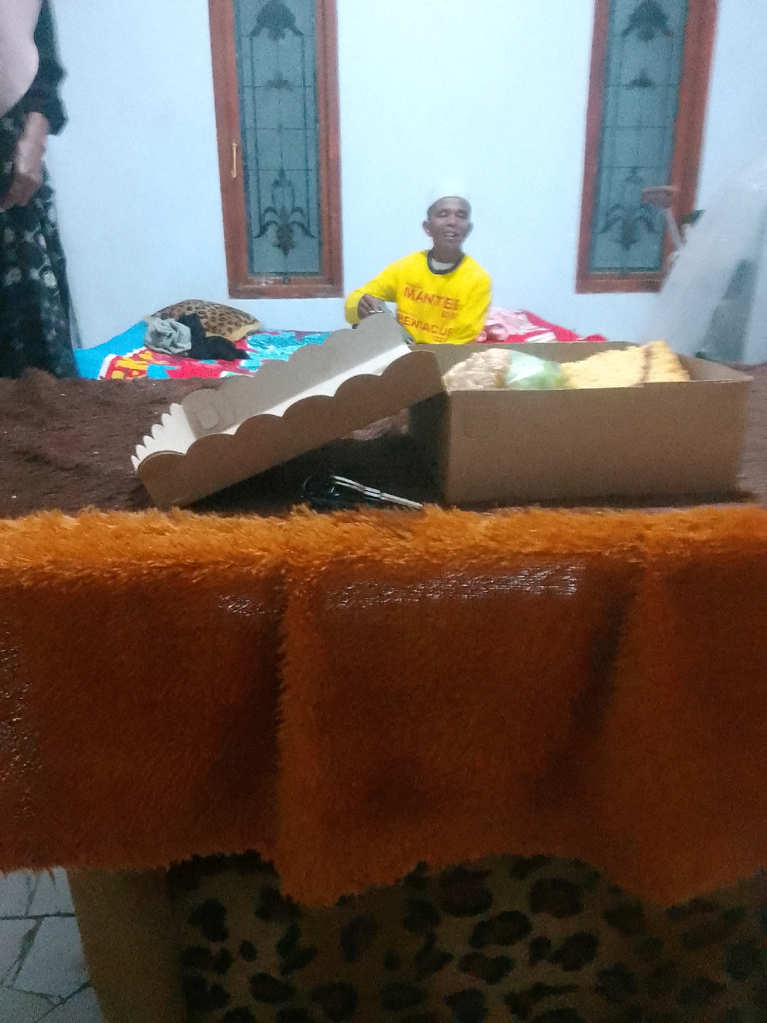 A young boy wearing a white cap and a yellow t-shirt with Indonesian text "MANTELL" and "RENACUS" printed on it is sitting indoors. He is looking at the camera and smiling. In front of him, there is a cardboard box containing food, possibly cake and snacks, partially covered by a white decorative lid. The boy is seated on what appears to be a bed or a carpeted surface. To the left of the boy, a portion of a person's arm and torso wearing a dark floral pattern garment is visible. The background shows two windows with wooden frames and decorative glass panes, and a white wall. The floor is covered with a dark brown carpet. In the foreground, there is an orange fuzzy surface, possibly a blanket or rug. The setting appears to be a room in a residential area in Krucil, Indonesia.