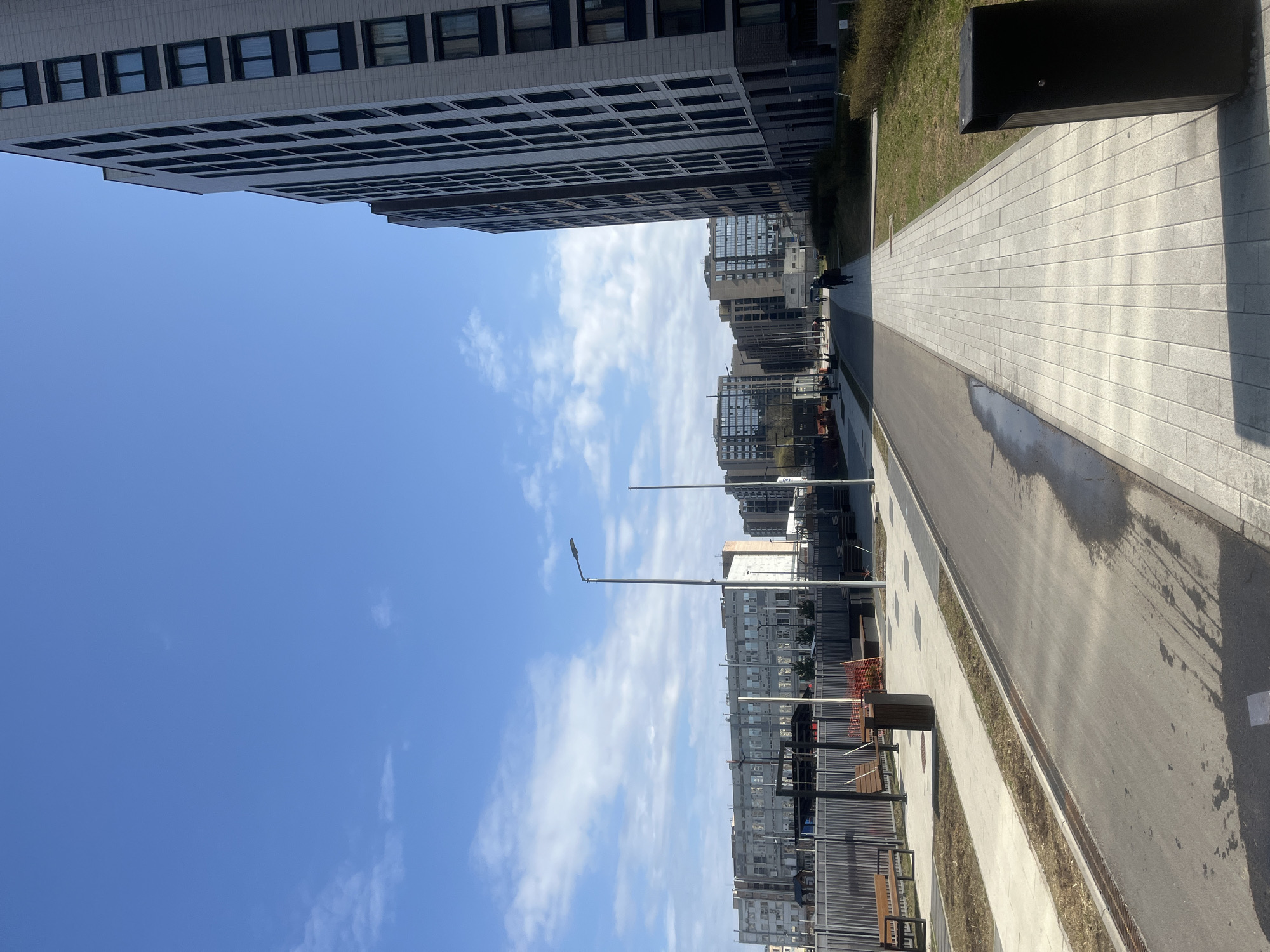 The image captures a street scene in Saint Petersburg, Russia. The view is oriented lengthwise down a pedestrian walkway flanked by modern buildings on both sides. The building on the left is tall and densely packed with windows. The sky above is blue with scattered white clouds. A row of streetlights lines the walkway. People are walking in the distance. On the right, a paved walkway runs alongside a road, with patches of grass and a black rectangular object, possibly a trash receptacle. The overall impression is of a clean, modern urban environment.