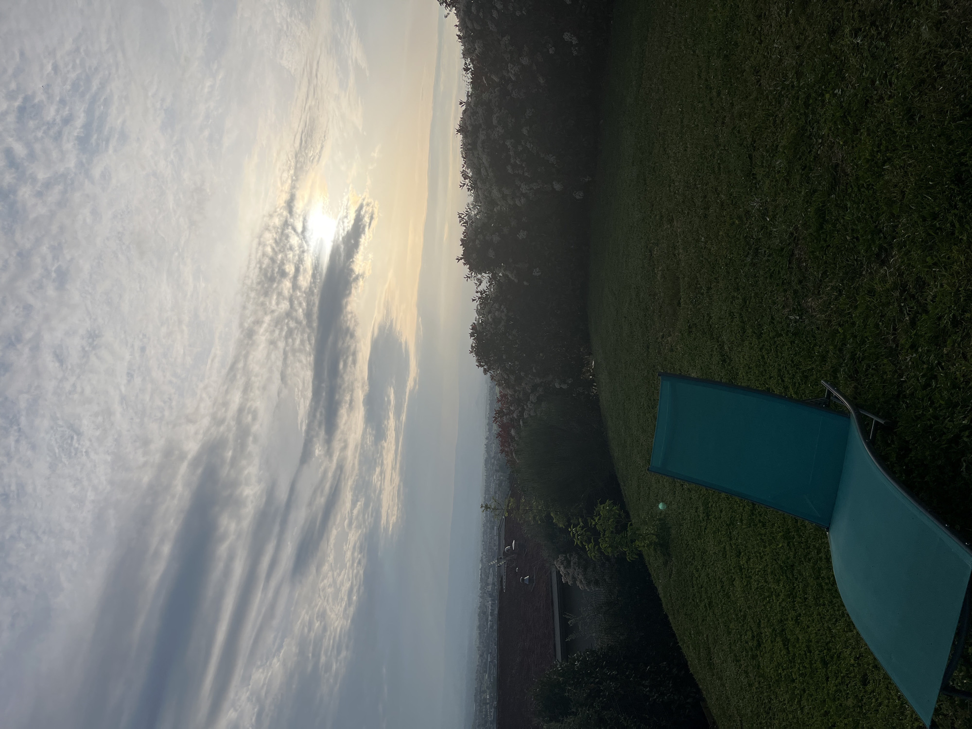 An outdoor scene, likely a garden or backyard, captured during either sunrise or sunset, overlooks a hazy distant landscape. The image is oriented such that the sky occupies the upper half, dominated by a mix of stratiform and cumulus clouds. The sun, partially obscured by clouds, is bright and low on the left side, casting a soft, warm light across the scene.
In the midground and foreground on the right, a vibrant green lawn is visible. A modern blue-green (teal) lounge chair with a dark frame rests on the grass, angled towards the left. A dense line of trees and bushes with light-colored blossoms runs along the right edge and forms a partial horizon line across the middle of the frame.
Beyond the treeline, a sprawling, hazy vista of a distant town or valley extends into the background. Some faint structures or rooftops can be discerned through the atmospheric haze. The overall setting suggests a tranquil, leisurely environment, consistent with a rural or semi-rural location such as Chambœuf, France. There are no people visible, nor any discernible text.