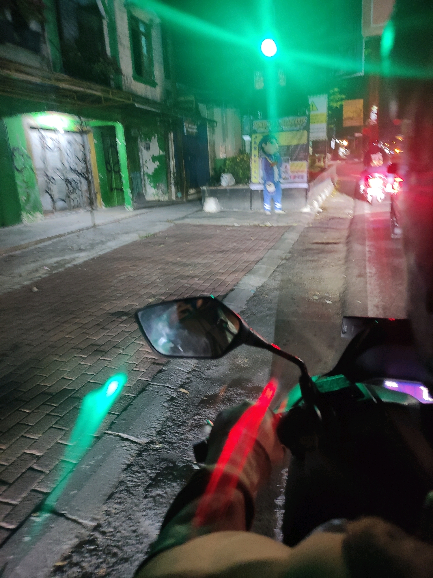 The image depicts a street scene at night, likely in Sleman, Indonesia, illuminated by green and red lights. The perspective is from a motorcycle, showing its mirror and handlebars. On the left, a building with green paint and graffiti is visible, with a sidewalk paved with brick. Further down the street, a person in a blue costume stands next to a sign. To the right, the taillights of vehicles create streaks of red light. The overall atmosphere is blurry due to motion and low light conditions.
