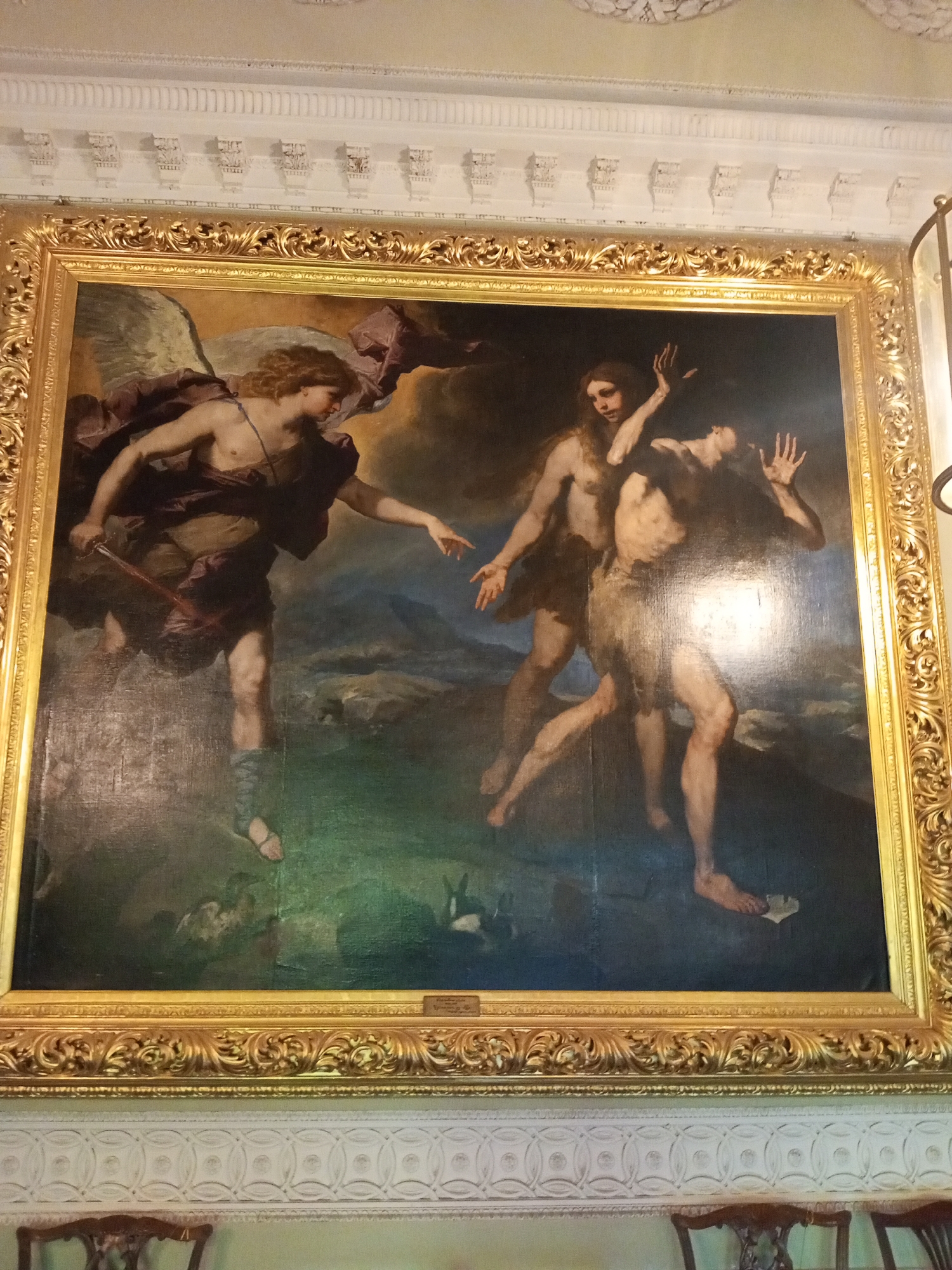 The image shows a framed oil painting depicting the expulsion of Adam and Eve from the Garden of Eden. An angel with wings and a sword points at Adam and Eve, who are recoiling in fear and shame. The background is a dark, stormy landscape. The painting is in a gilded frame and is hanging on a wall with elaborate white molding. The picture was taken in the Palace of Gatchina located in Gatchina, Russia.