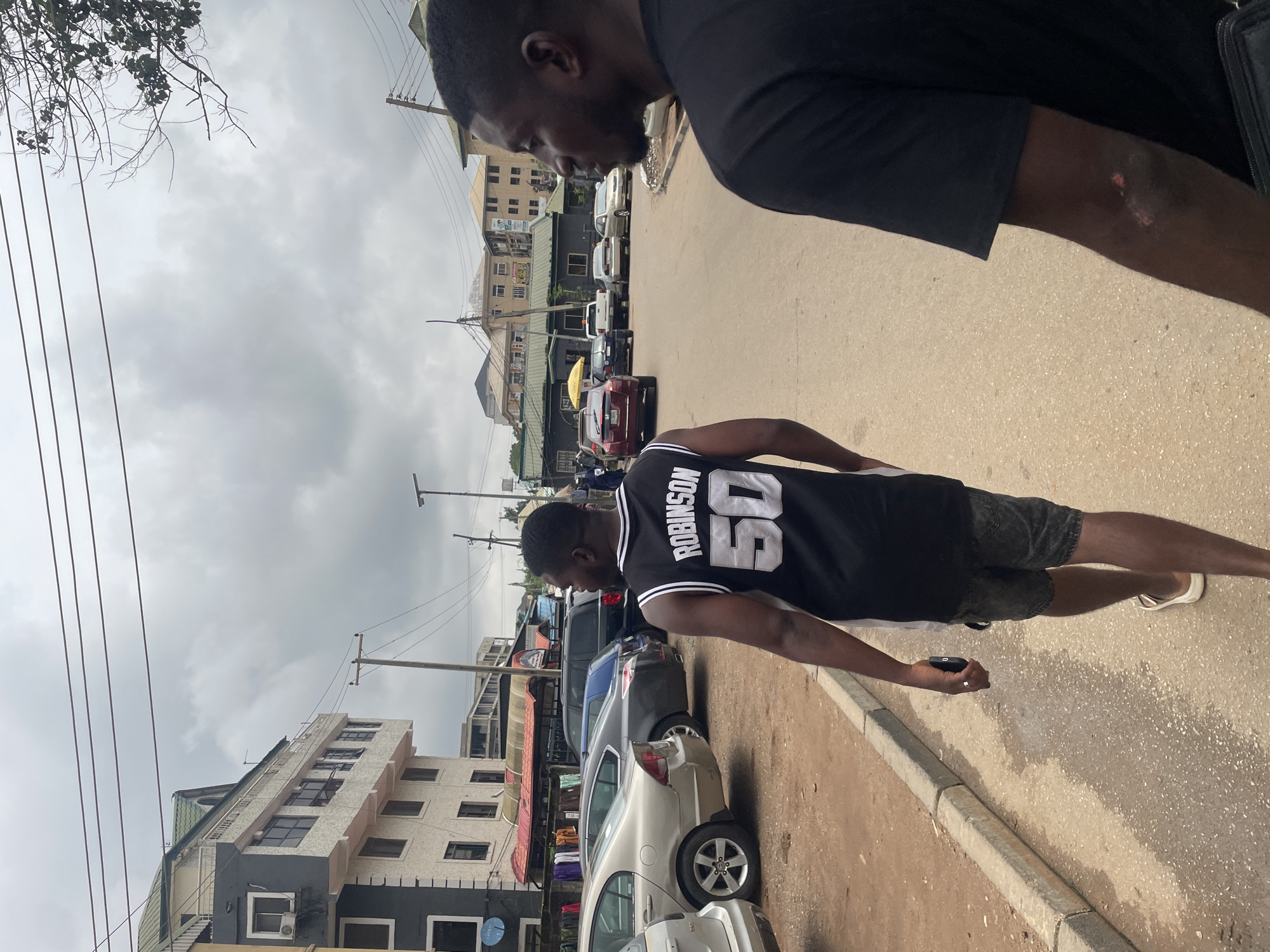 Two young Black men are walking on a paved street in Abuja, Nigeria. The man in the foreground on the left is wearing a black basketball jersey with "ROBINSON 50" printed in white on the back, and gray shorts. The man on the right is wearing a black t-shirt and is slightly out of focus. The street is lined with parked cars, including a silver sedan, a dark blue SUV, and other vehicles in the background. Buildings with multiple stories, some with balconies and air conditioning units, are visible on the left side of the street. The sky is overcast with scattered clouds. Power lines run overhead, supported by utility poles. The ground appears slightly wet in places.