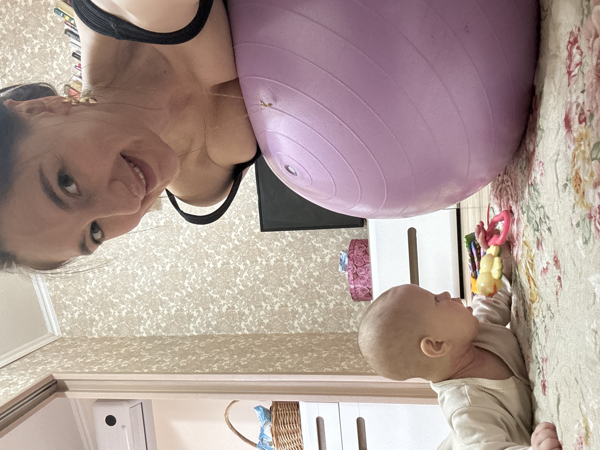 A rotated eye-level shot shows a woman with dark hair smiling at the camera, leaning her head against a large, purple exercise ball. To the right of the ball, a baby lies on a floral patterned surface, reaching for a colorful toy. Behind the woman and the ball is a TV screen mounted on a wall with a patterned wallpaper. The location appears to be indoors, potentially a home, but there is no indication this is in Sochi, Russia.