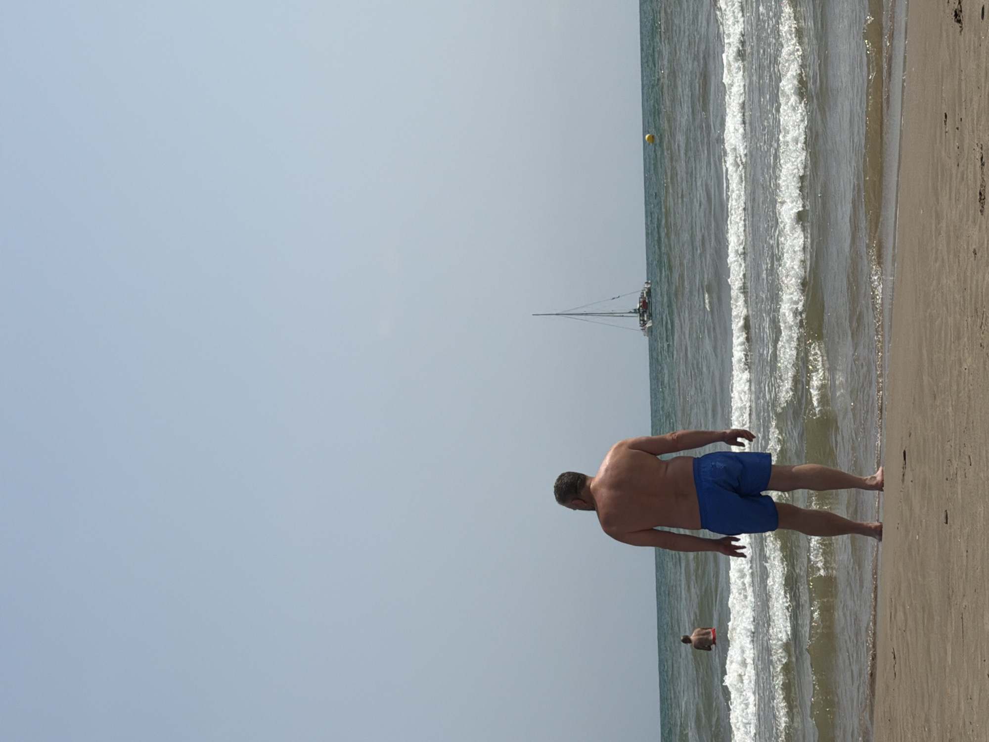 A full shot from behind shows a man standing on a sandy beach facing the ocean under a clear, pale blue sky. He is wearing blue swim trunks and is shirtless, with his back to the camera. His skin appears tanned, and he has short, dark hair. In the middle ground, waves crash on the shore. Further out in the ocean, a sailboat with a tall mast is visible, along with a yellow buoy. To the left of the sailboat, a person is standing in the water, wearing red swim trunks. The water is a greenish-blue color. The sand in the foreground is light brown with some small pebbles. The image appears to be taken on a sunny day.