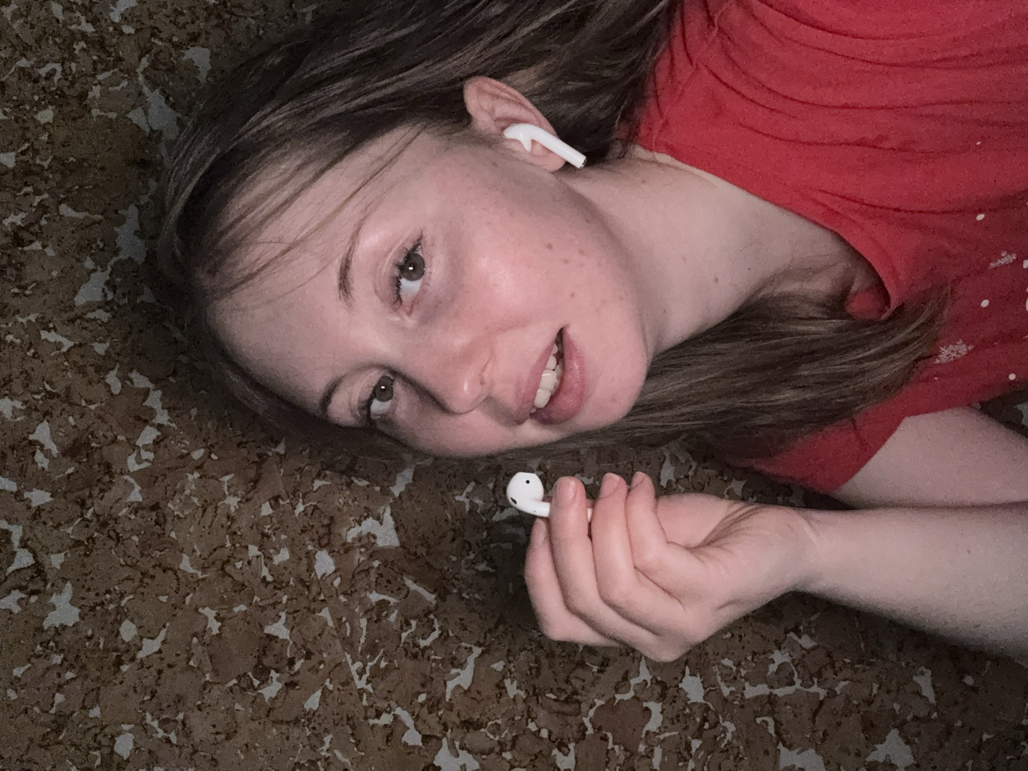 A fair-skinned woman with light brown hair is lying on a surface resembling cork. She wears a red shirt and has one white wireless earbud in her right ear. She holds another identical earbud in her right hand. The location appears to be indoors, with no immediate indicators of a specific city or place. The environment suggests a domestic setting with cork-like flooring or a similar textured surface.