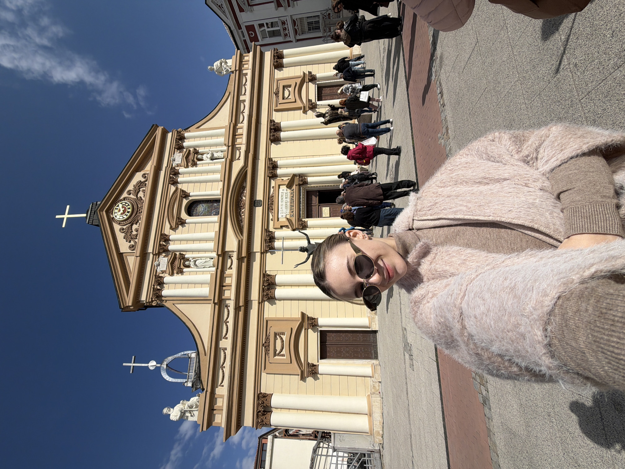 A woman in a brown sweater and a fuzzy pink shawl is taking a selfie in front of the Cathedral of St. Louis in Plovdiv, Bulgaria. The church is a large, ornate building with yellow walls, brown trim, and white columns. A group of people are walking towards the front entrance. The sky is blue with some clouds. The photo appears to be taken at ground level.