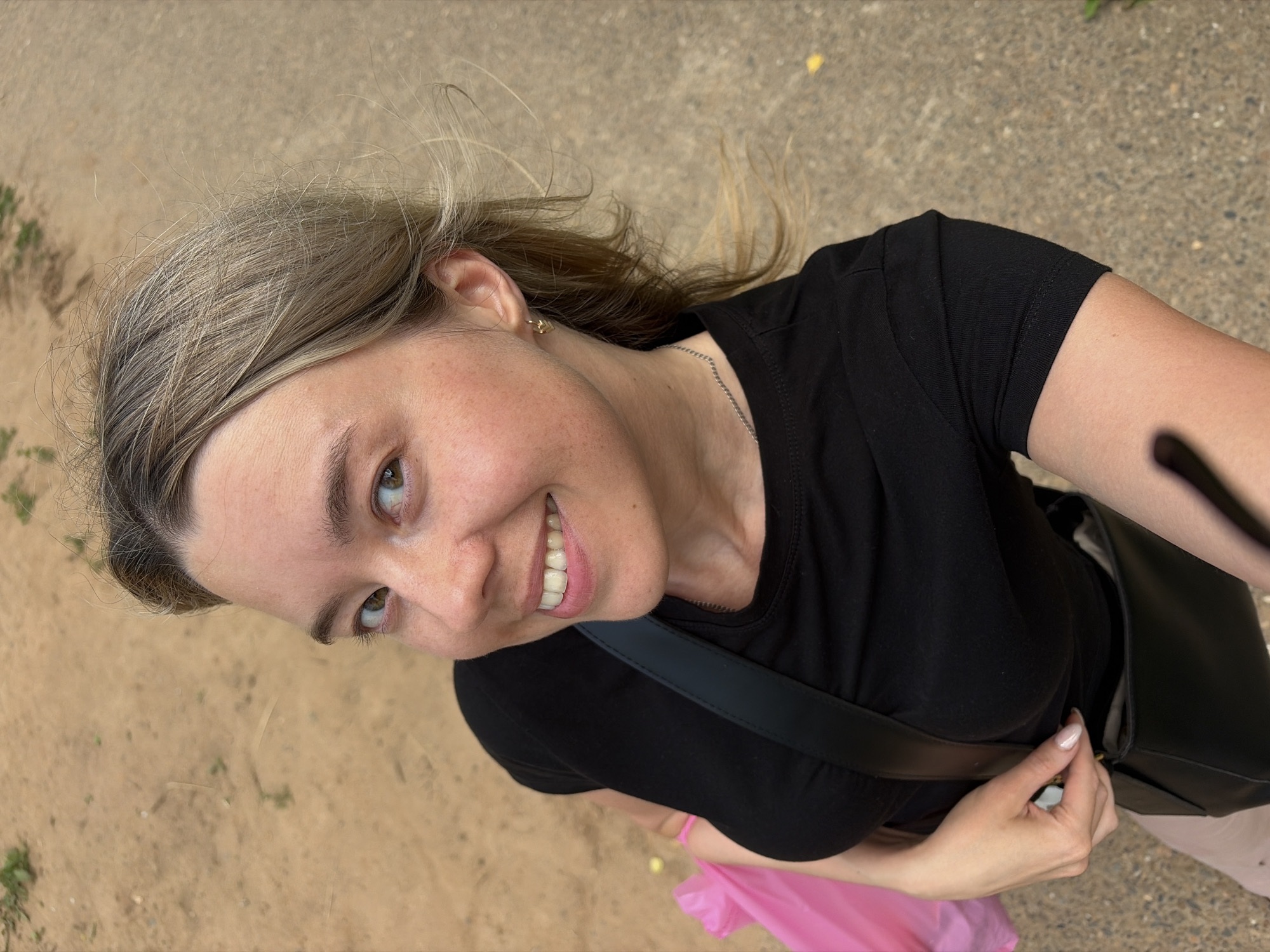 A young woman with blonde hair and green eyes smiles at the camera. She is wearing a black t-shirt, a silver necklace, and a small gold earring. A black purse with a strap is slung across her body, and she holds a pink bag in her left hand. The background is a sandy path, possibly in a park or outdoor area. The image appears to be a selfie, taken from a high angle. The location is identified as Chaikovsky, Russia.