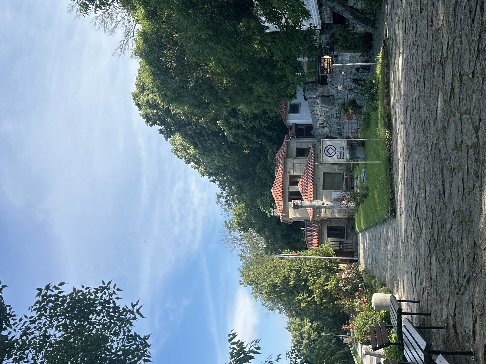 The image shows a stone pathway leading to a complex of stone buildings with red tiled roofs, situated on a hillside covered in dense green trees. In the foreground, there are two wooden benches facing the pathway. To the left of the pathway, there is a patch of grass with red roses and a cylindrical stone object. To the right of the pathway, there is a green lawn with rose bushes and a stone wall. A sign with the text "World Heritage Convention" is visible on the left side of the pathway. A yellow directional sign with text in Cyrillic is posted on the right side of the pathway. A Bulgarian flag is planted near the buildings. The sky is blue with scattered white clouds. The scene appears to be an outdoor tourist attraction or historical site.
