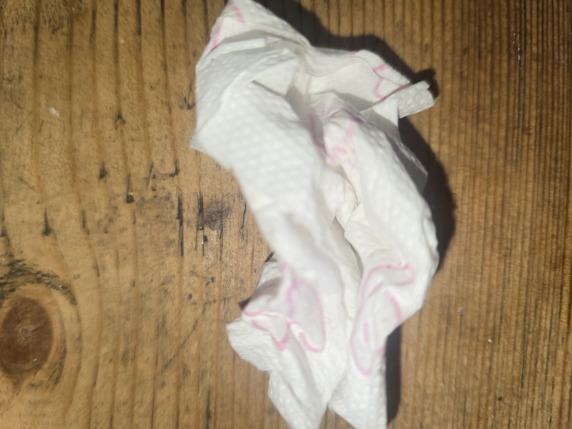 A crumpled white paper napkin with pink trim lies on a wooden surface. The wooden surface is made of planks with visible grain and knots.