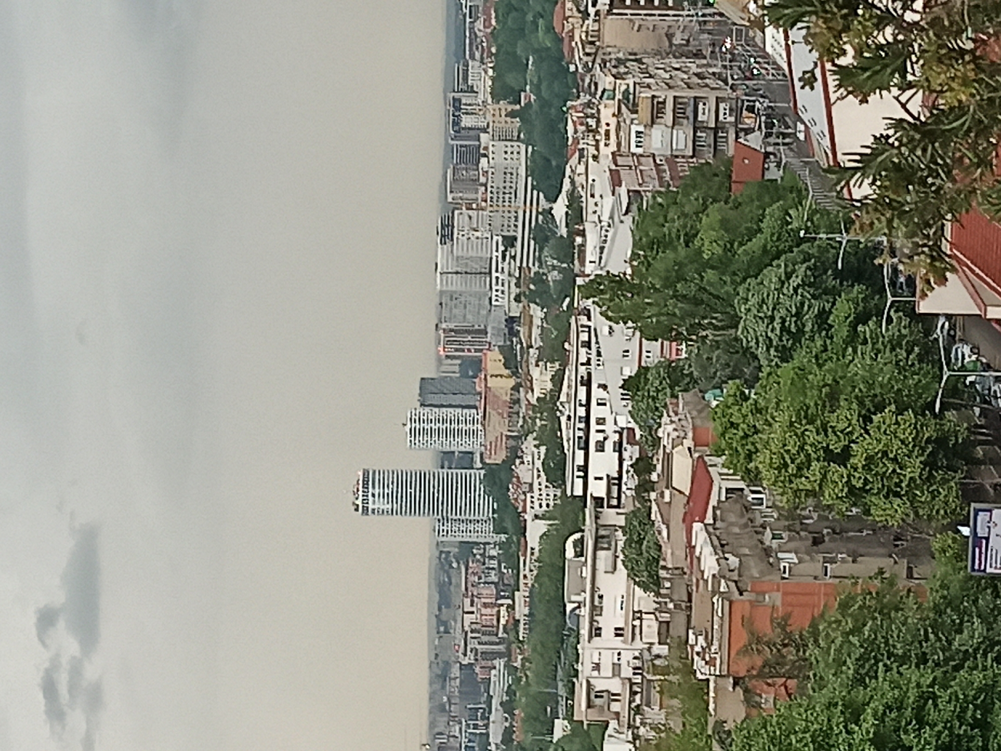 This image depicts a cityscape in Vozdovac Urban Municipality, Serbia. The foreground shows residential buildings with red rooftops interspersed with lush green trees. In the distance, modern high-rise buildings and other urban structures are visible, partially obscured by haze or smog. The sky is overcast. The perspective is likely from an elevated vantage point. Street lights and other urban fixtures are present, and a sign is visible in the lower right corner. No people are visible in the shot.