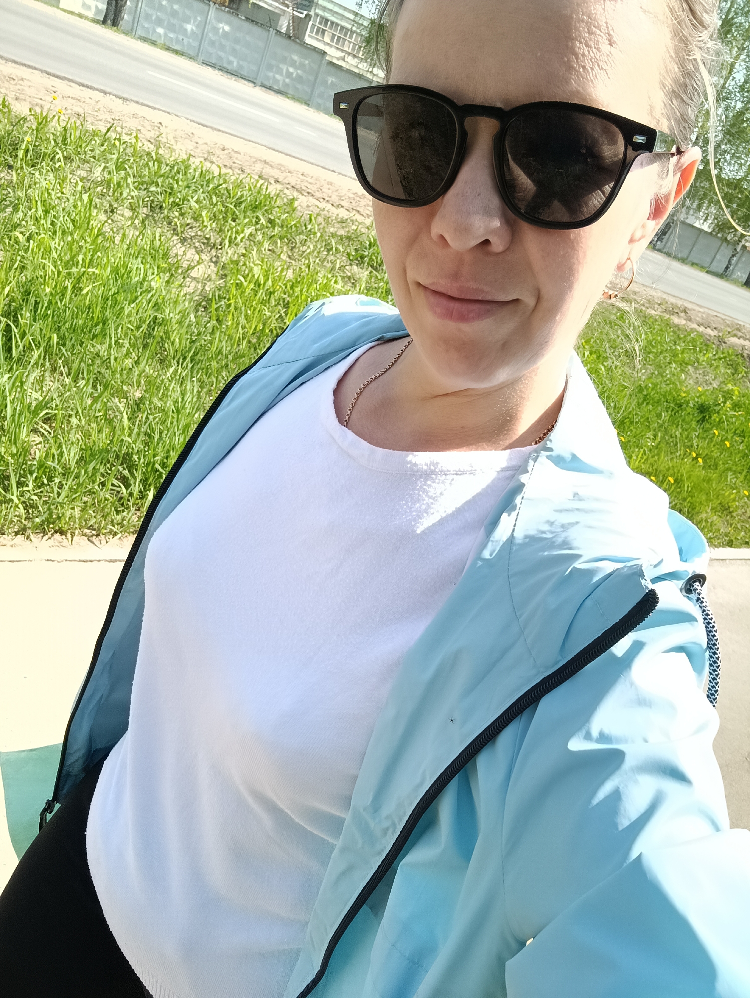 A woman is taking a selfie outdoors. She is wearing sunglasses, a white t-shirt, and a light blue jacket. She is also wearing a gold necklace and earrings. The background includes green grass, a sidewalk, a road, and a fence. The image was taken in the city of Chaikovsky, Russia.