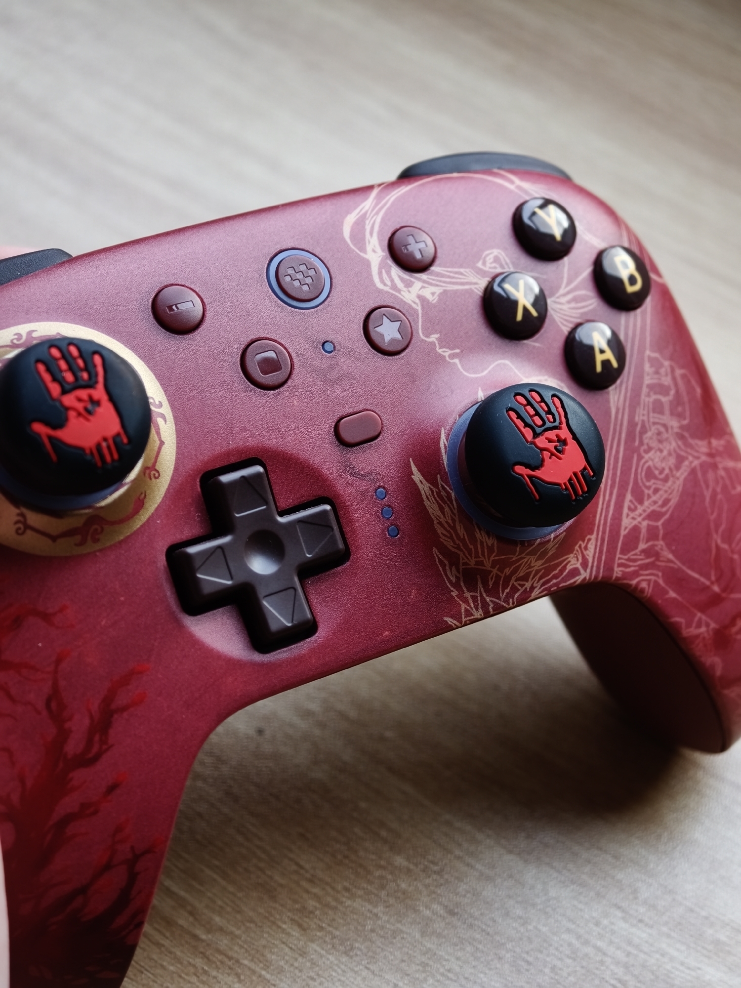 A close-up, angled shot shows a red video game controller with intricate gold line art designs. The controller has several buttons and joysticks. Two of the joysticks are covered with black caps featuring a red, bloody handprint design. The controller rests on a light brown, wood-grain surface. The image does not provide any information about its location in Đà Nẵng, Vietnam.