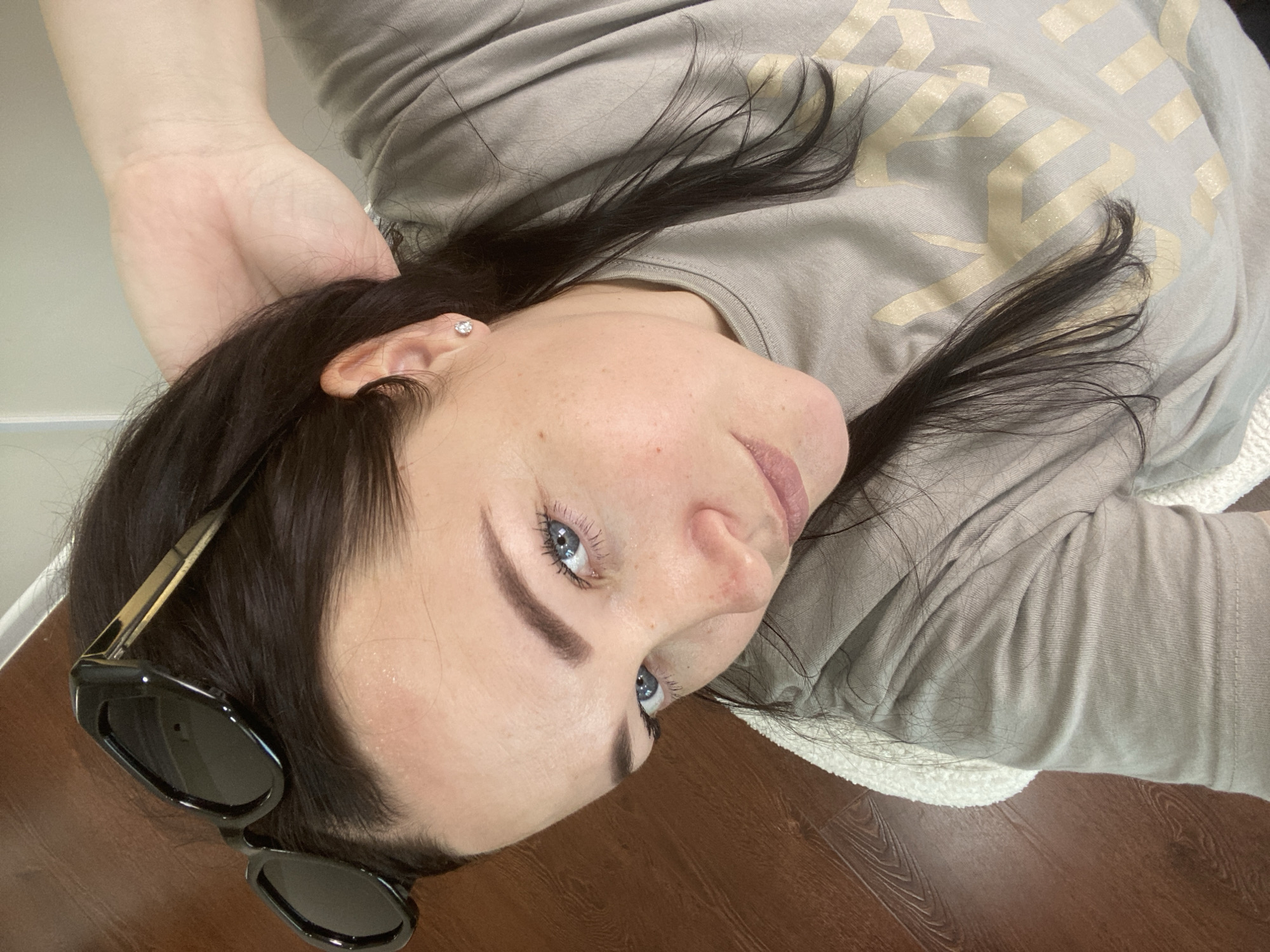 A woman with dark hair and blue eyes is lying down, wearing a grey t-shirt and black sunglasses. Her left hand is raised towards her head. The background includes a wooden floor and a white, fluffy rug. There is no indication of the city of Sochi, Russia.