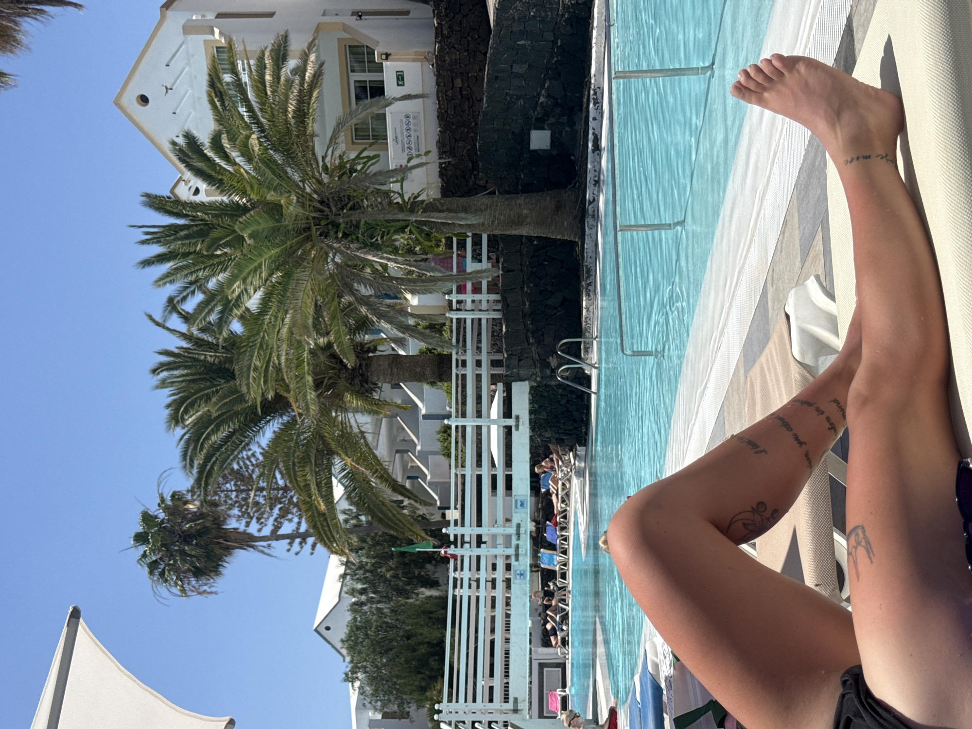 The image shows a person relaxing by a pool at a resort in Puerto Del Carmen, Spain. The person's legs, with tattoos, are visible on a white lounge chair. The pool is light blue and surrounded by a white fence and lounge chairs with other people relaxing on them. Palm trees are visible in the background, along with white resort buildings and a clear blue sky. The resort appears to be next to a black lava rock wall.