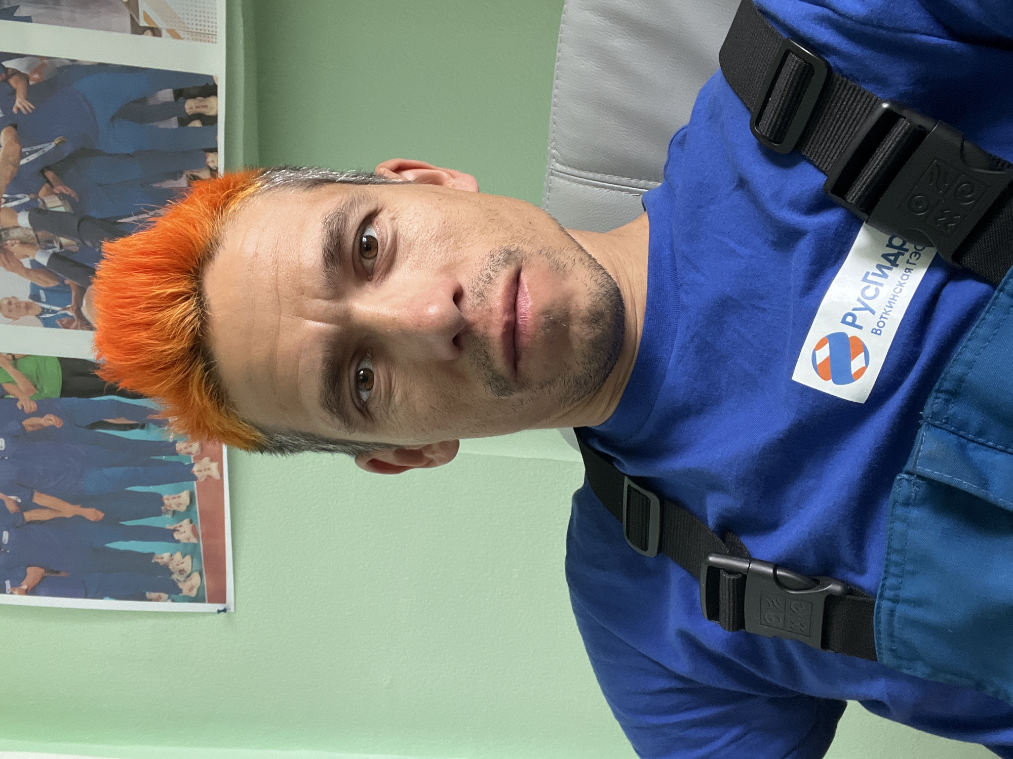 A man with bright orange, spiky hair on top and darker hair on the sides looks directly at the camera with a neutral to slightly serious expression. He has light brown eyes and noticeable facial stubble. He is wearing a blue short-sleeved t-shirt and what appears to be blue work overalls or a similar garment, evidenced by a black strap with a plastic buckle worn over his right shoulder.
The setting is indoors, likely an office or workplace common area, with a plain light green wall visible in the background. A grey upholstered chair back is partially visible behind his head. To his left, a poster is attached to the wall, depicting several people standing in blue uniforms.
A prominent white logo patch is affixed to the man's blue t-shirt. The logo features a circular graphic composed of orange, white, and blue segments, resembling an energy symbol or turbine. Above and below this graphic, Cyrillic text is visible. The upper text reads "РУСГИДРО" (RusHydro), and the lower text reads "Воткинская ГЭС" (Votkinskaya HPP), indicating the Votkinskaya Hydroelectric Power Plant. This detail, combined with the location context of Chaikovsky, Russia, confirms the setting as a facility associated with the RusHydro company and the Votkinskaya HPP. The man's attire suggests he is an employee at this location.