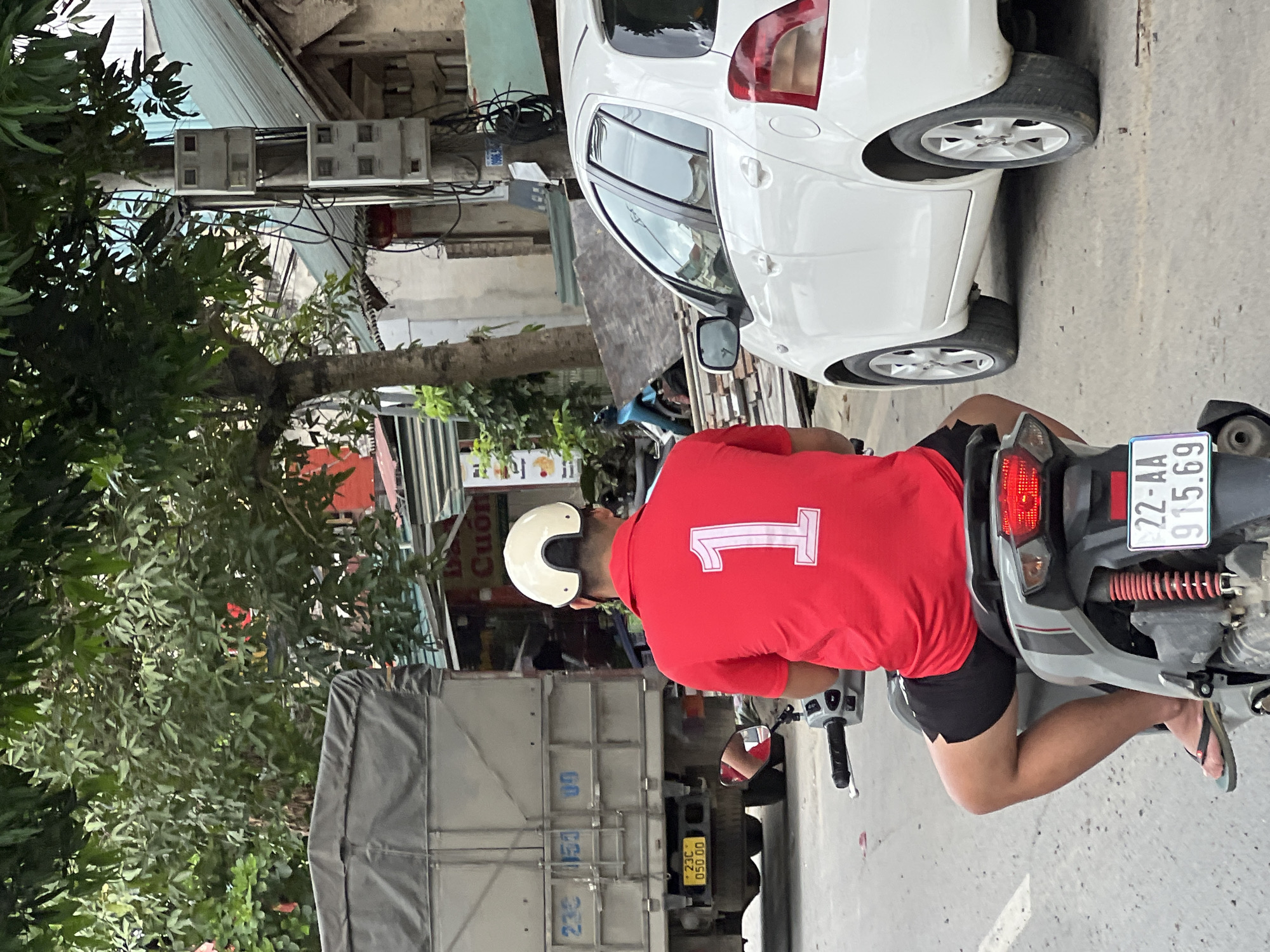 In Hà Giang 2 Ward, Vietnam, an individual wearing a red polo shirt with a white number "1" emblem and black shorts is seated on a scooter. They are also wearing a white helmet. The scooter is gray with red accents. The individual's left leg is visible, and they are wearing a black flip-flop. The scooter's license plate reads "22-AA 915.69". In the background, a large gray truck is parked, with the partial license plate "23C 050.00" visible. A white car is parked to the right of the scooter. Trees and utility poles with multiple meters are also present in the environment. The ground is paved.
