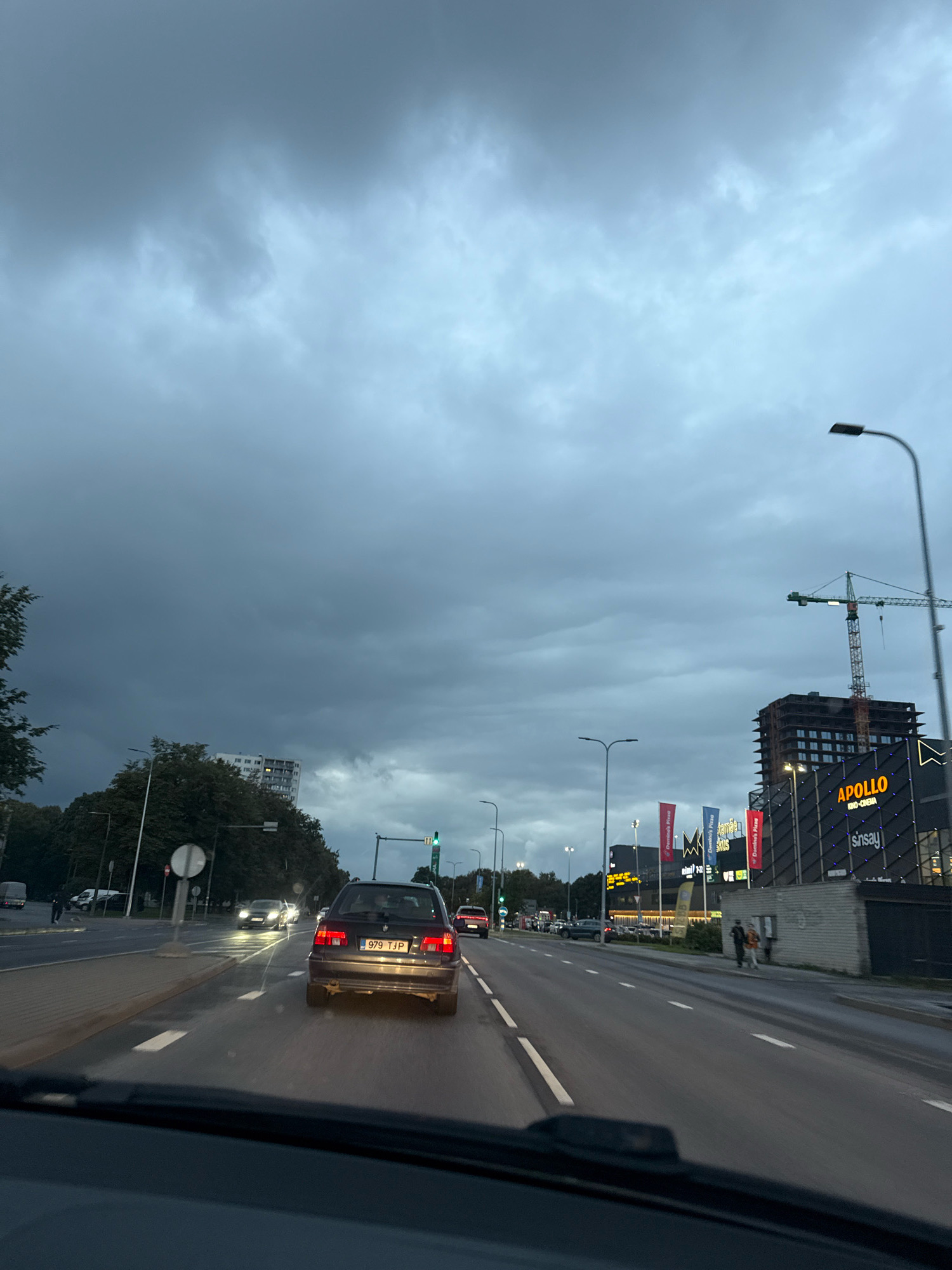 The image captures a view from a vehicle driving on a multi-lane road in Tallinn, Estonia, under a heavily overcast sky, suggesting impending rain or a storm. The road surface appears wet, with lane markings visible. Several cars are on the road, with headlights on, indicating low visibility. On the right side of the road, a modern building complex is visible, featuring signage for "APOLLO" and "Sinsay." A construction crane is also present near the building, indicating ongoing development. The left side of the road features trees and a tall apartment building in the distance. The overall atmosphere is somber due to the dark clouds and the muted lighting conditions.