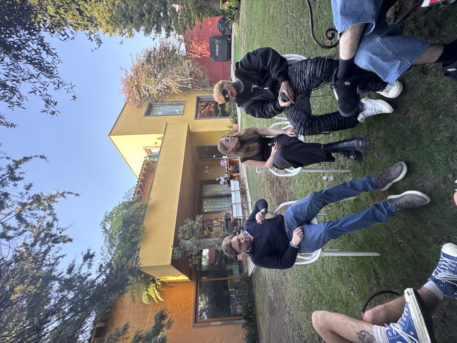 A group gathers on a grassy lawn outside a modern yellow building. Three people sit in white chairs: a person in a black jacket and patterned pants, a woman in black dress and boots, and a man in a blue sweater and jeans. Parts of others are visible, including denim-clad legs and blue sneakers. A grill stands in the background, partially obscured by a flowering tree. The sky is clear and blue.
