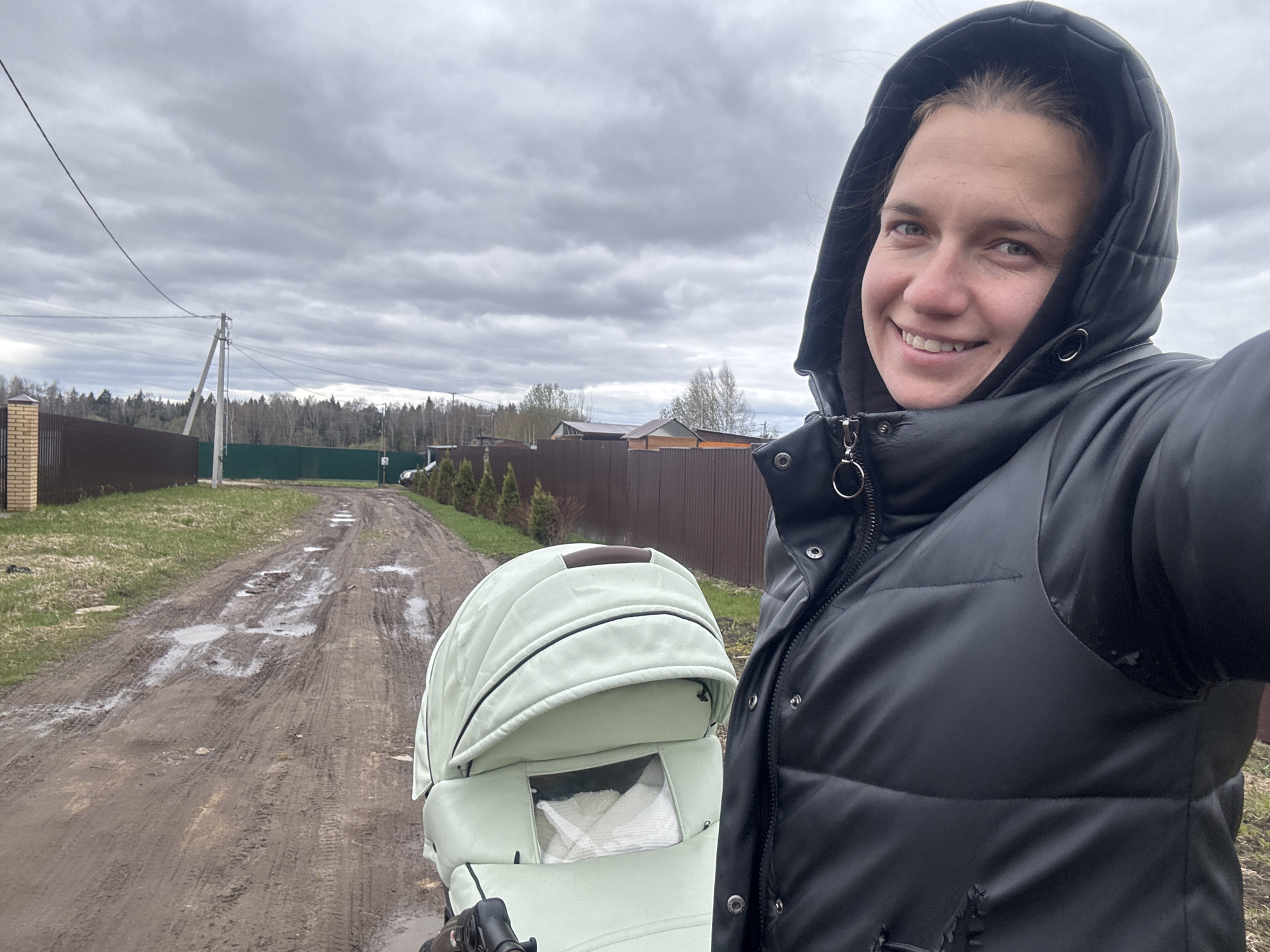 A smiling woman takes a selfie while pushing a light green baby carriage on a muddy, unpaved road in Раздолье, Russia. The woman, wearing a black hooded, puffy jacket, is positioned on the right side of the frame, looking towards the camera with a cheerful expression. The baby carriage, a pram with a light green canopy and a small viewing window revealing a white blanket inside, is prominent in the lower-left and central parts of the image.
The scene is set outdoors under an overcast sky filled with heavy gray clouds, suggesting a gloomy or recently rainy day. The dirt road is visibly muddy with numerous puddles, reflecting the dull sky and indicating wet conditions. Along the left side of the road, there's a strip of grass, followed by a brick pillar and a dark brown fence. Utility poles and power lines are visible further back, extending into the distant tree line. On the right, a row of evergreen shrubs lines a dark green fence, with several houses visible beyond it in the background. The overall impression is of an everyday walk in a rural or semi-rural environment during an overcast period. There is no visible text in the image.