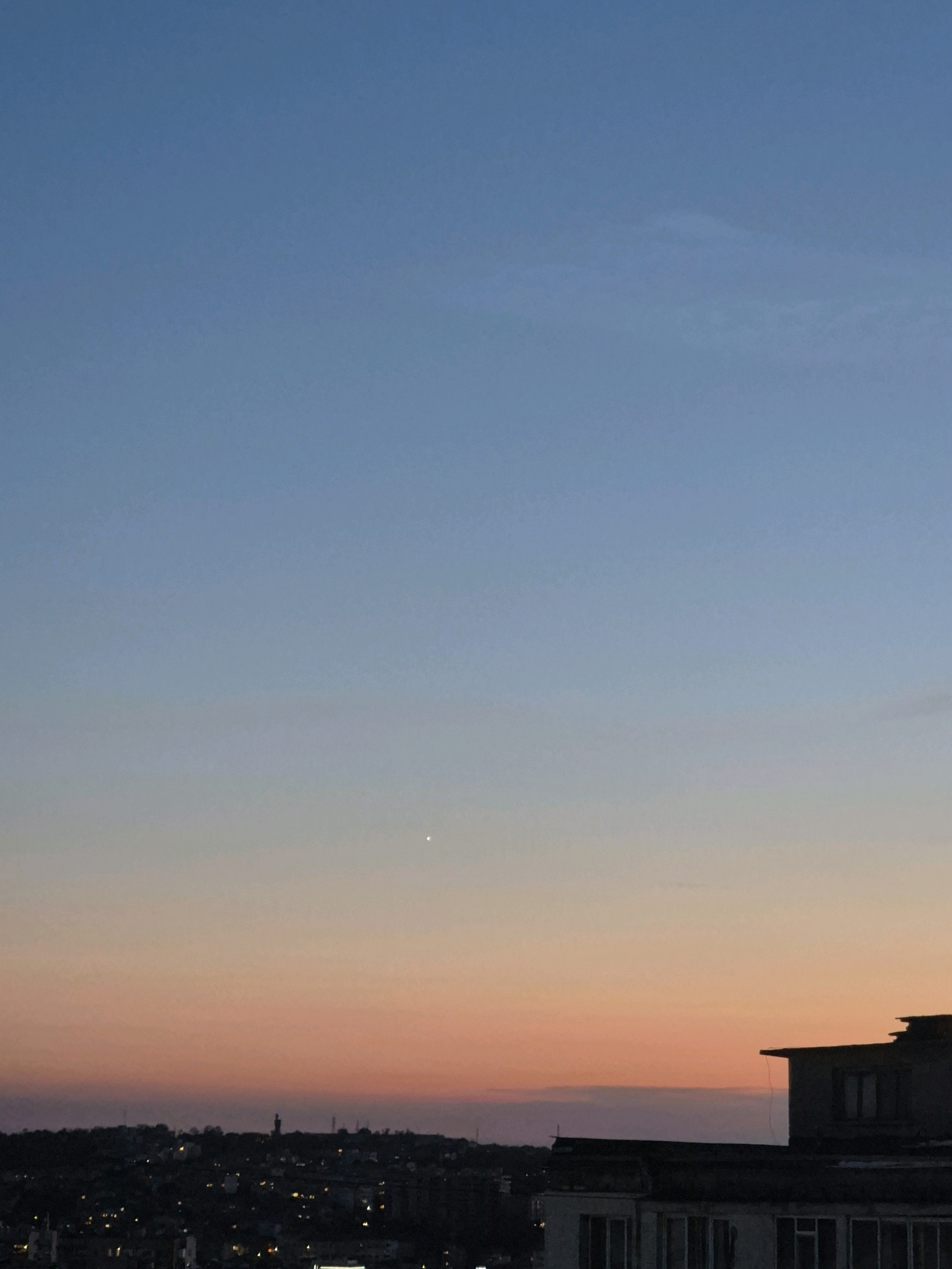 A cityscape is visible under a gradient sky transitioning from deep blue at the top to soft orange and pink hues near the horizon. A solitary bright star or planet is visible in the upper portion of the sky. Silhouettes of buildings, some with illuminated windows, form the skyline. In the foreground, the roofline of a dark, angular building is prominent, featuring a window and some debris on its top. The overall scene suggests dusk or dawn.
