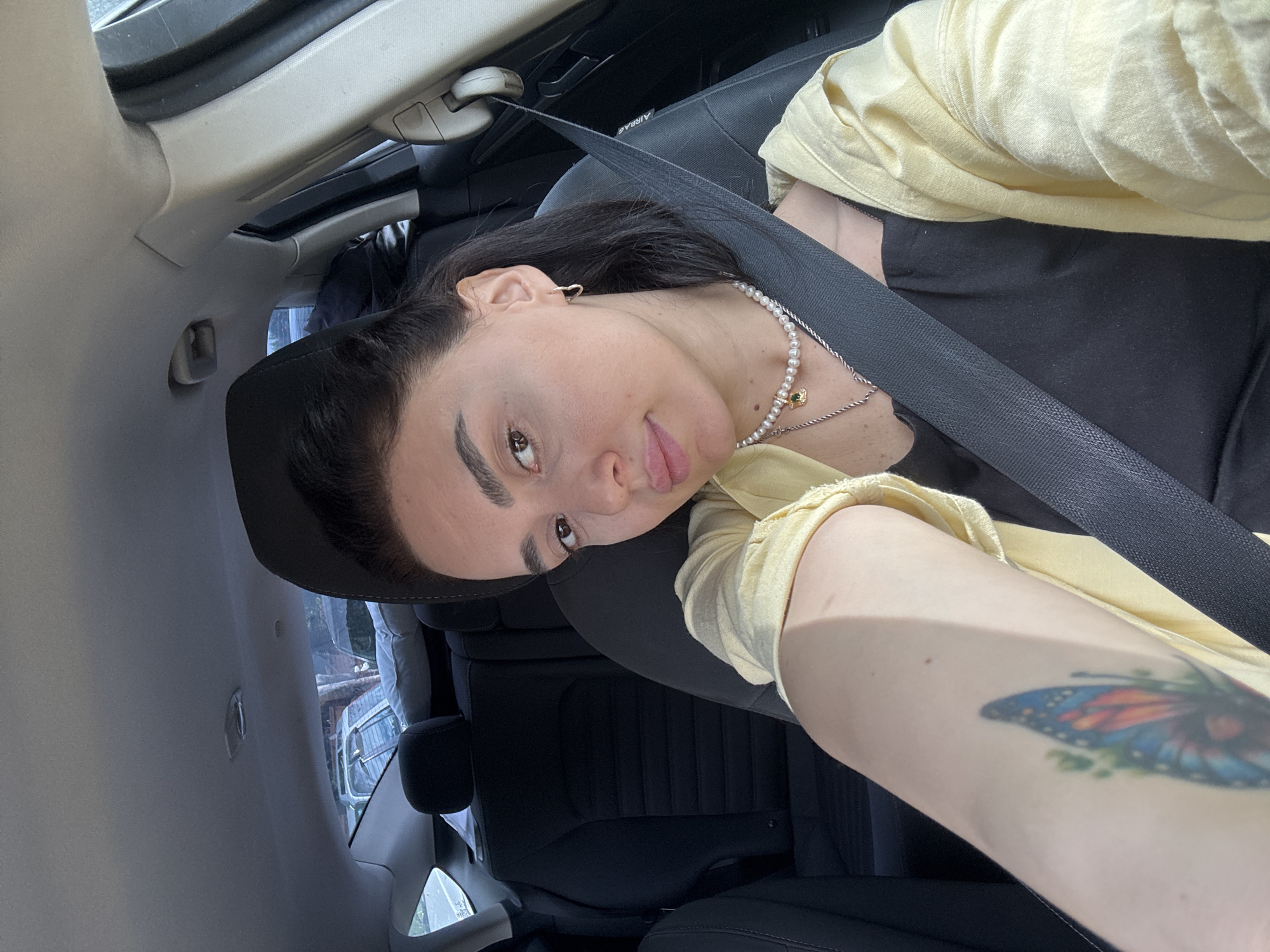 A person with dark hair and light skin is inside a car, wearing a black top and a yellow cardigan, along with a seatbelt. The person has a butterfly tattoo on their left arm. The car's interior is visible, including the roof, seats, and a glimpse of the outside environment. The location is Tbilisi, Georgia, visible through the car window as buildings and other vehicles.
