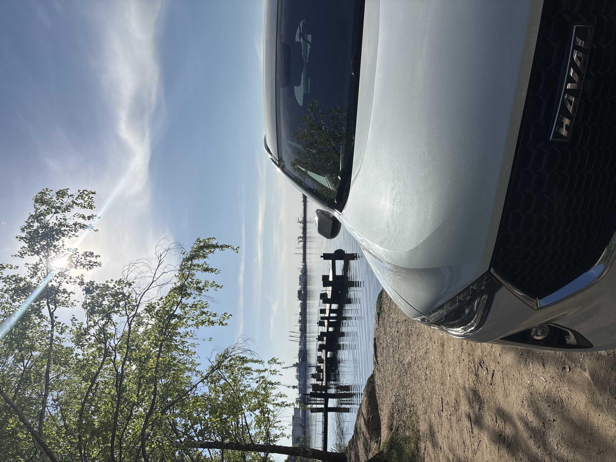 The image shows a white car, identified by the "Haval" emblem, parked on a dirt area next to a body of water. Pilings are visible in the water, extending into the distance. Across the water, the skyline shows industrial structures, including cranes. A tree with green leaves is present in the left foreground. The sky is blue with wispy clouds, and the sun is shining through the tree. The location is Chaikovsky, Russia, based on the style of buildings on the far shore of the water.