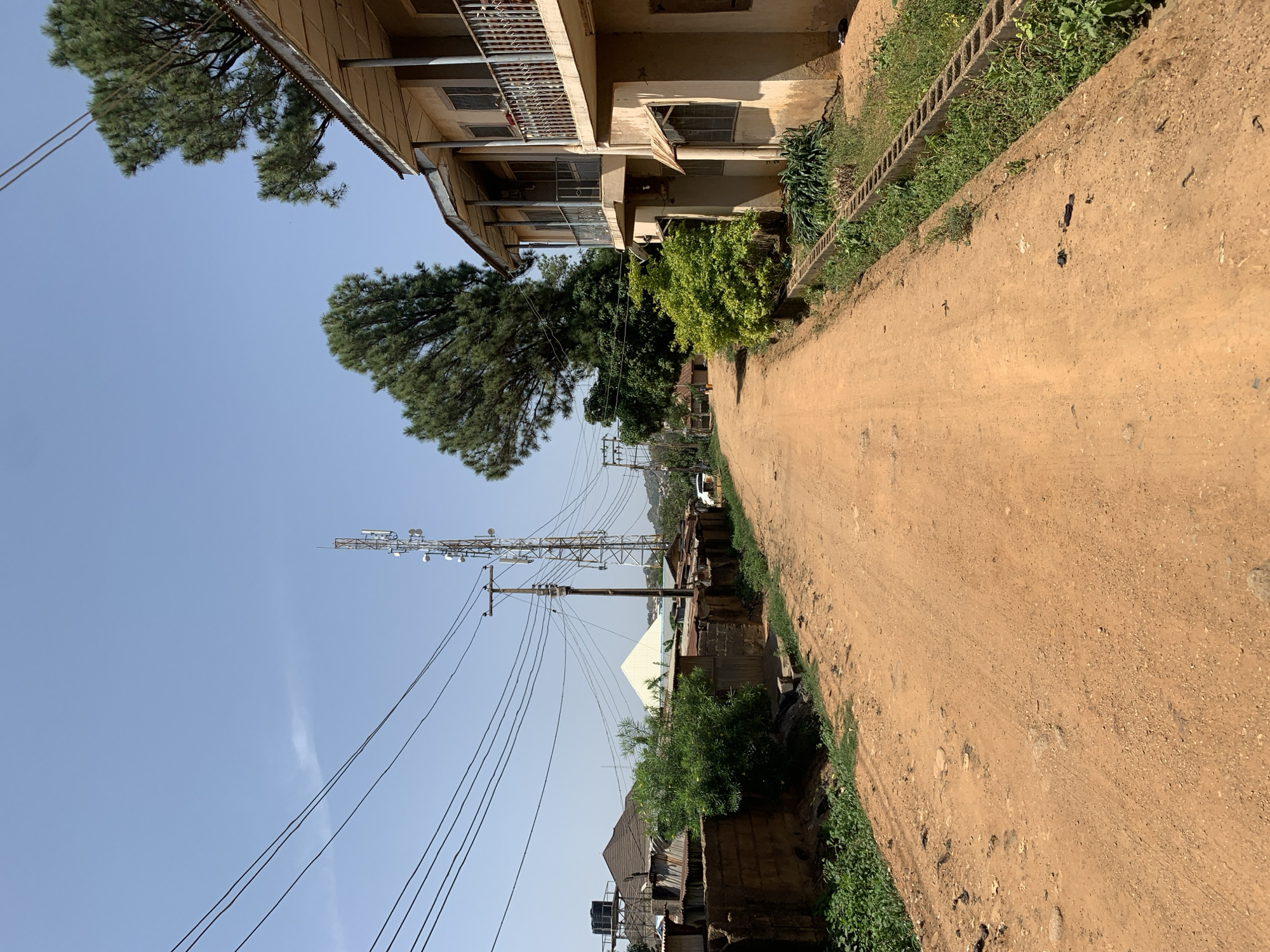 The image depicts a dirt road in Rafin Par, Nigeria. The road is bordered by buildings on both sides. On the left, there is a two-story building with balconies. On the right, there is a lower structure. A tall communications tower is visible in the distance. Power lines run parallel to the road. The sky is clear and blue. The light suggests it is daytime. There are no people visible in the frame.