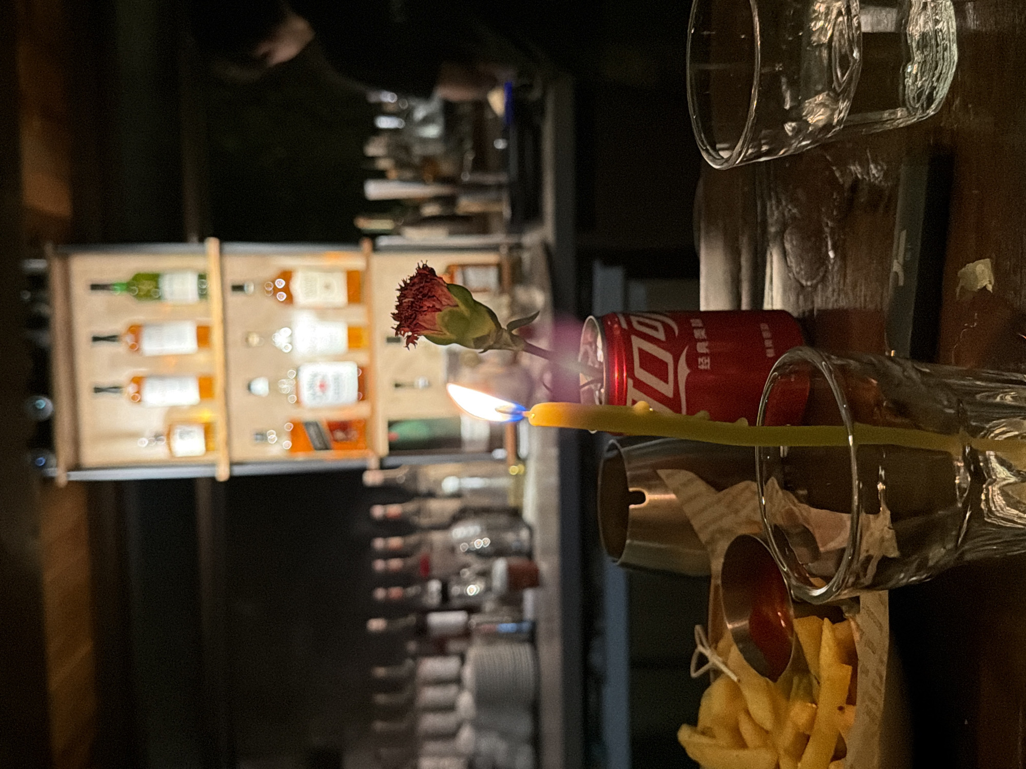 The image captures a dimly lit interior scene, likely a bar or restaurant, set in Longgang District, China. In the foreground, a lit yellow candle stands in a glass. To the right of the candle is a red Coca-Cola can, which holds a single dark red flower. In front of these objects is a plate of french fries with a small dish of dipping sauce. To the right of the frame, a partially filled glass of clear liquid sits on a wooden table. In the background, shelves are stocked with various bottles of alcohol. A person, seen from the back, is visible in the far right, standing behind a counter with bottles and glasses. The overall ambiance is intimate and possibly romantic, with the candle flame providing the primary light source.