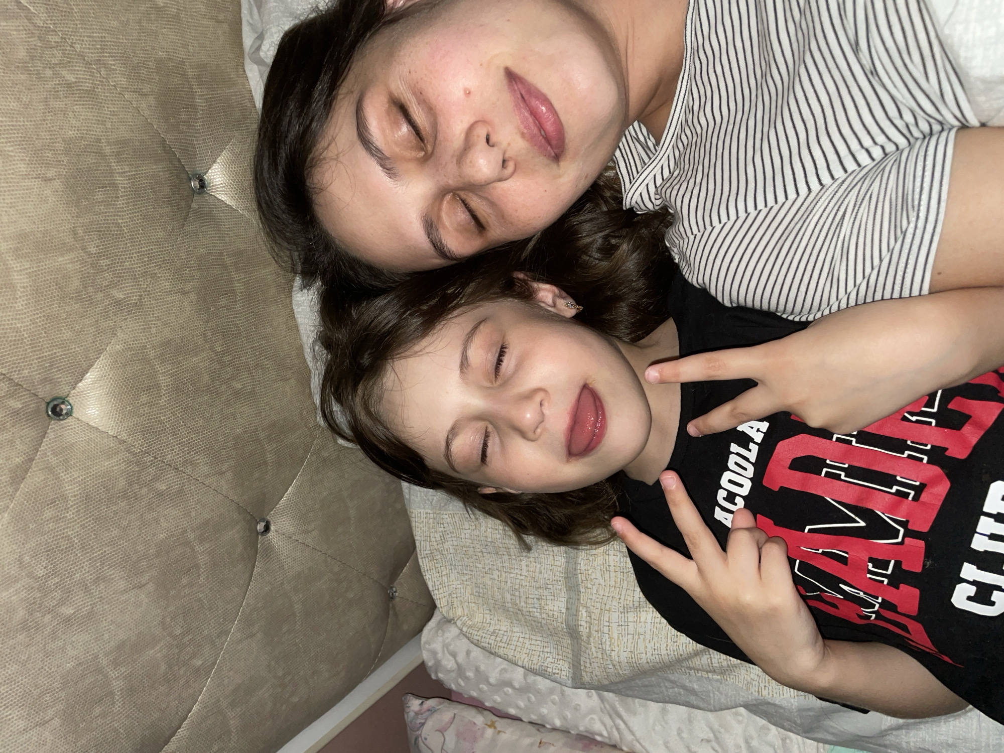 Two individuals, presumably a woman and a young girl, are lying down side-by-side on a bed. Both have their eyes closed. The woman is wearing a striped top. The young girl is wearing a black t-shirt with red and white lettering. The girl is making a "peace" sign with both hands. The headboard of the bed has a textured, quilted design. The image was not taken in Izhevsk, Russia. The location of this image is impossible to determine from the available information.