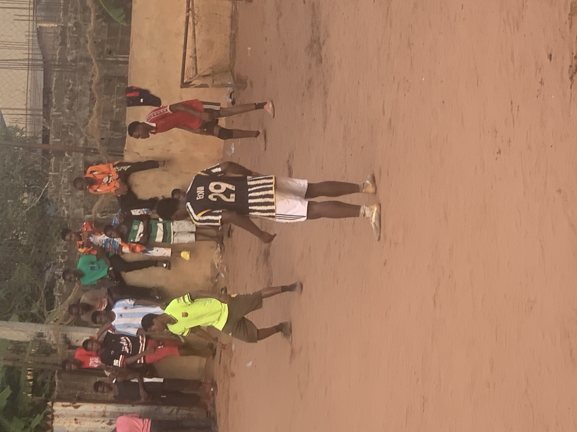 The image depicts a lively gathering of men and boys on a reddish-brown dirt field, likely for a football (soccer) game or practice in Onitsha, Nigeria. The scene is illuminated by the soft, warm light of what appears to be late afternoon or early evening.
Numerous individuals are scattered across the dusty ground, many dressed in various football jerseys and shorts. In the midground, a prominent figure stands with his back to the viewer, wearing a black and white striped jersey featuring the number "29" and the name "BONI" across the shoulders. He is also wearing white shorts and light-colored athletic shoes. Nearby, another person in an orange long-sleeved shirt is bent over, while a player in a red jersey and black shorts is also visible.
Further to the left, a cluster of individuals can be seen. One person is wearing a bright yellow-green shirt and dark shorts, appearing to be in motion. Other jerseys visible include a light blue and white striped shirt (resembling the Argentina national team kit), black and white striped patterns, and plain red. Many participants are young, and their attire suggests active play.
The setting features unpaved, dry, reddish-brown earth. In the background, there are hints of structures that appear to be partially constructed or unrendered buildings with exposed rebar and brickwork, alongside some sparse green vegetation. A metal frame, possibly part of a goalpost, is faintly discernible in the upper right background. A dark bag rests near a wall. The overall impression is of an informal sporting event or training session taking place in a community setting.
