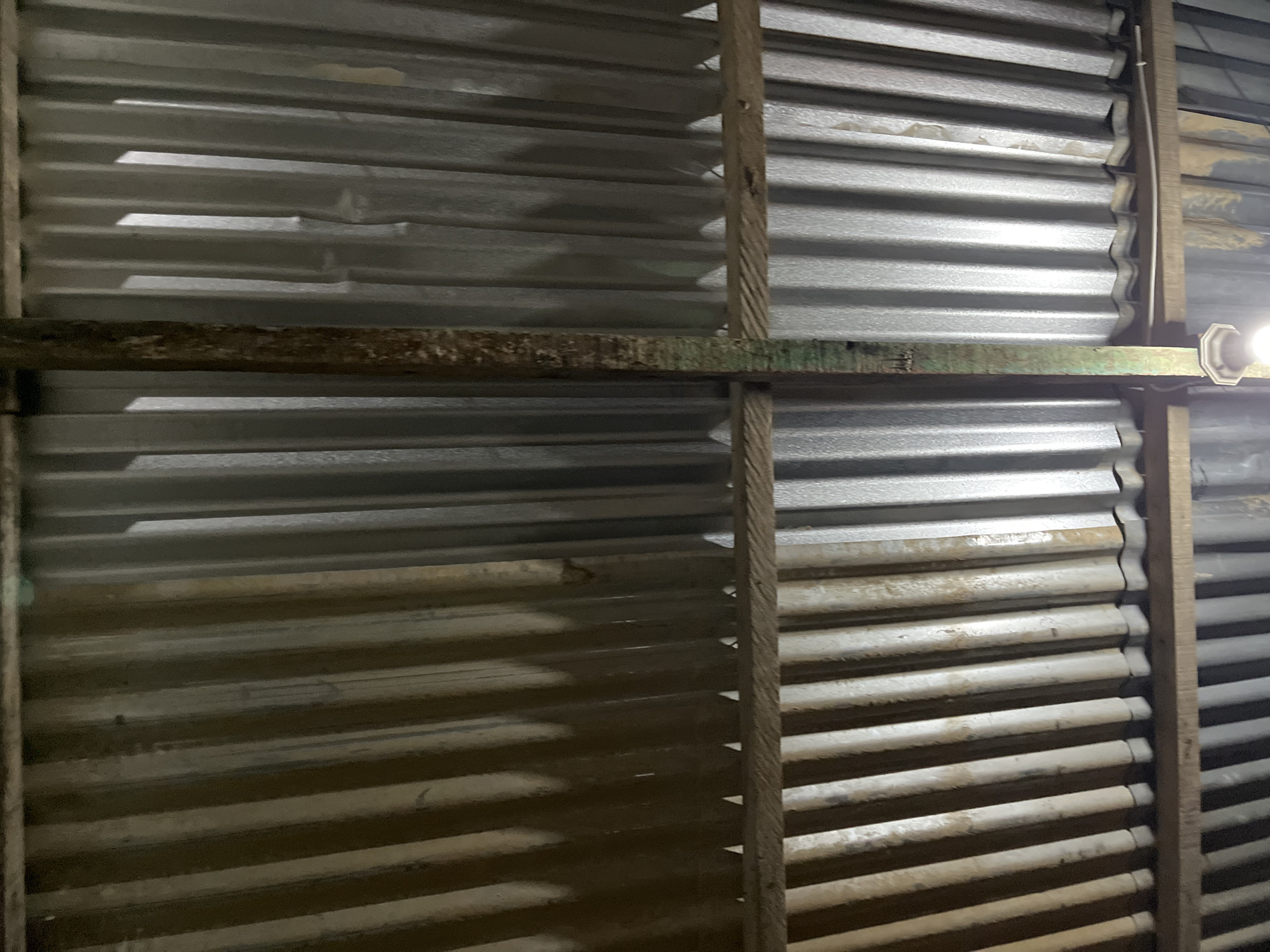 The image shows the corrugated tin roof of a structure, viewed from below. The roof is supported by wooden beams and a vertical wooden post. The tin sheeting is weathered and shows signs of rust and dirt. The lighting suggests it is either dusk or the structure is poorly lit from within. The environment appears to be a rustic or utilitarian building, possibly in a rural or semi-urban area. Without additional information, pinpointing the exact location in Benuaraja, Indonesia is not possible. The image does not contain any people, actions, or interactions.