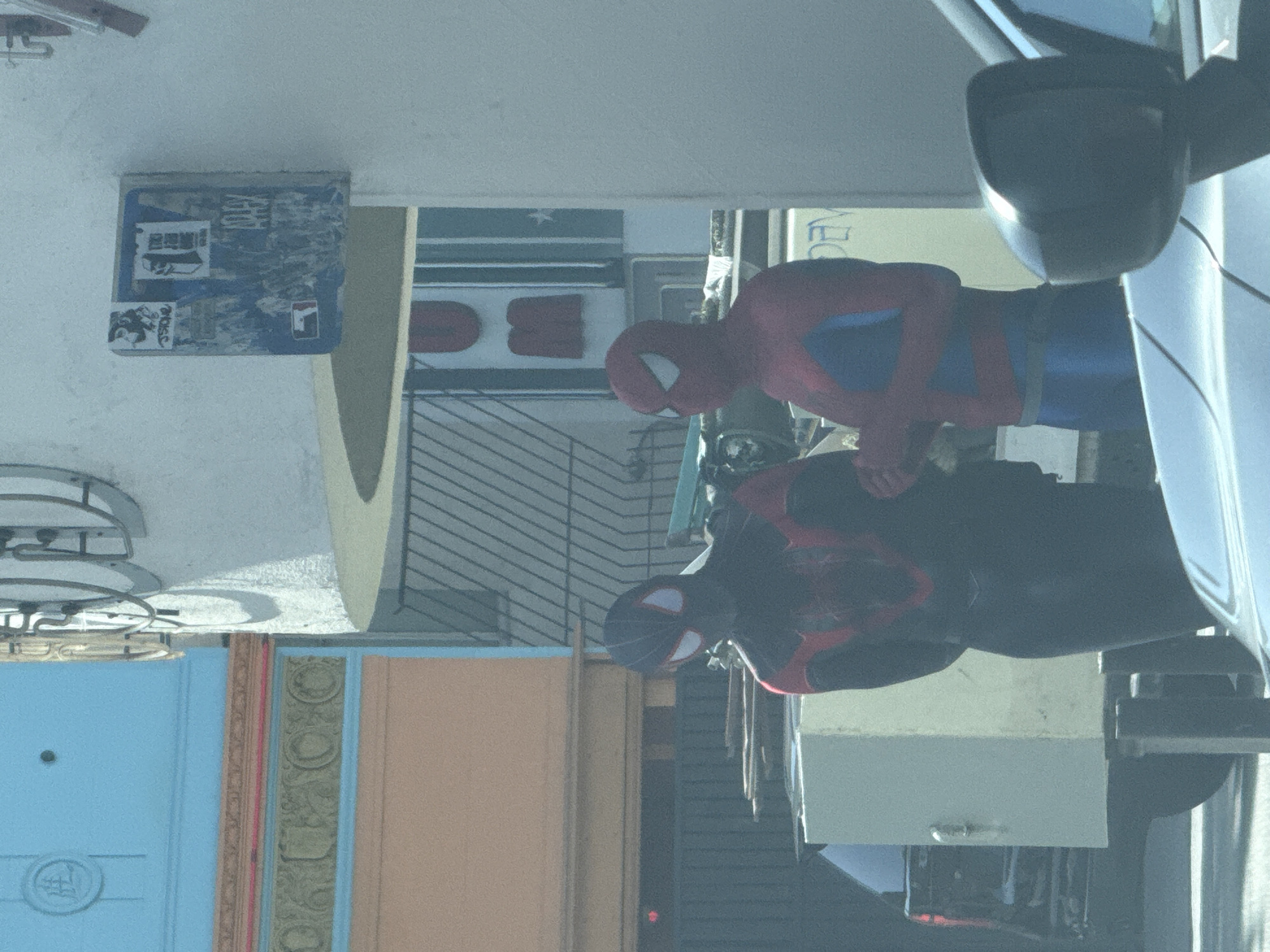 The image, when rotated 90 degrees clockwise, displays an outdoor urban scene featuring two individuals dressed as Spider-Man and Miles Morales. The classic red and blue Spider-Man stands on the right, while Miles Morales in his black and red suit is positioned to the left and slightly in front. Both characters appear to be standing, possibly posing, in what looks like a street or sidewalk environment.
In the immediate foreground on the right, a portion of a white vehicle, including its side mirror, is visible. The background consists of building facades. On the upper left, a blue, box-like structure is attached to a wall, covered with several stickers. One clearly shows the "FILA" logo, and another appears to have Asian script. Behind the classic Spider-Man, a sign with partially visible red lettering shows "DE". Below this, red block-shaped elements are present. To the far left, multiple doors are visible, including a light blue one with a circular emblem and a reddish-brown door with decorative carvings. The scene is brightly illuminated, indicating daytime. The setting suggests a lively public space, likely in Los Angeles, United States of America.