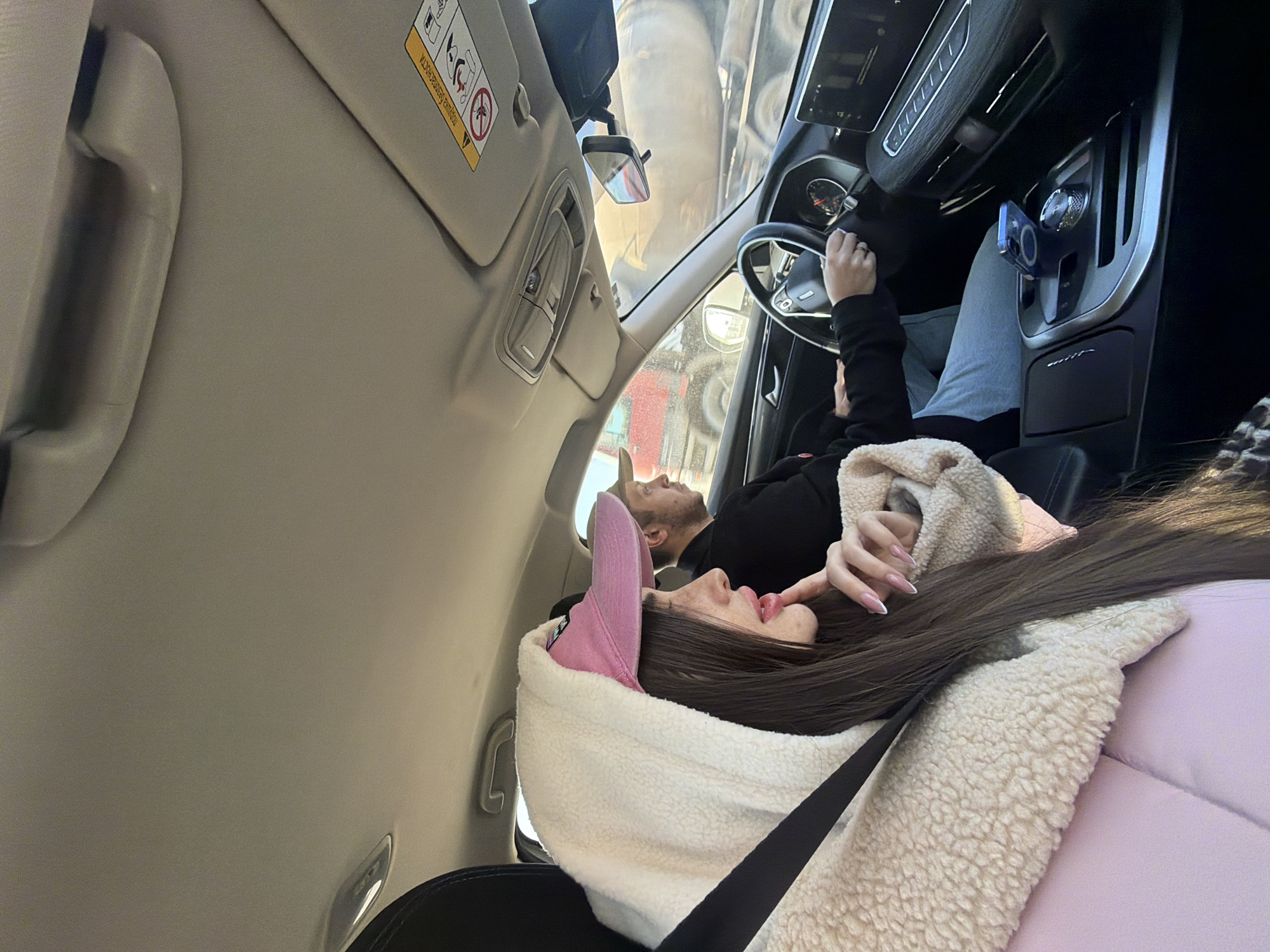 The scene depicts the interior of a moving vehicle from the perspective of a rear passenger, looking forward. Two individuals are present: a female passenger in the front seat on the right, and a male driver in the front seat on the left.
The female passenger has long, dark hair and is wearing a cream-colored, textured hooded jacket over a pink garment or seat cushion. She also wears a pink baseball cap with a dark logo. A black seatbelt crosses her chest. Her right hand, adorned with manicured nails featuring white tips, is positioned near her chin/lower lip. She is looking forward and slightly to her right.
The male driver, partially obscured, wears a dark top and a light-colored baseball cap. His hands are on the steering wheel, which bears the logo of a vehicle manufacturer (possibly Haval). He is focused forward.
The vehicle interior is modern, featuring a digital display panel on the dashboard to the right of the steering wheel and a rotary gear selector on the center console. A blue smartphone with a circular magnetic attachment is resting on the center console. The car's ceiling is light-colored. A sun visor on the passenger side displays a warning label in Cyrillic script, indicative of an airbag system.
Outside the passenger window and windshield, a large industrial truck, likely a cement mixer, is visible in motion, partially obscured. The truck features light-colored bodywork with red components. The external environment suggests daytime conditions.
Based on the visual evidence, specifically the Cyrillic warning label, the general location is a Russian-speaking region. However, there are no specific landmarks or identifiers within the image to definitively pinpoint the city as Saint Petersburg, Russia.