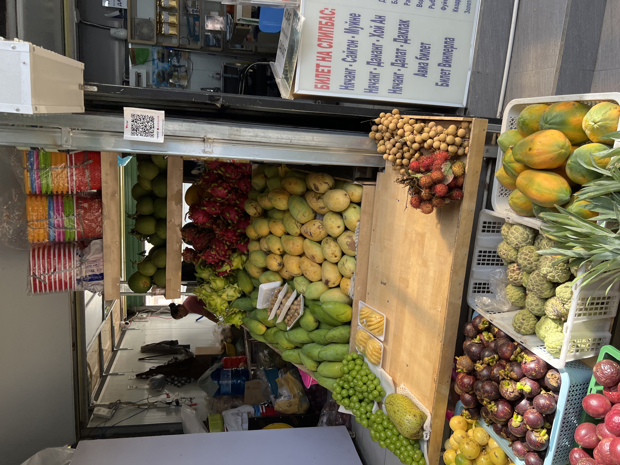 The image shows an outdoor fruit stand in Nha Trang, Vietnam. The stand is filled with a variety of fresh fruits, including dragon fruit, mangoes, grapes, custard apples, mangosteens, papayas, rambutan, and longan. There are also some packaged sliced pineapple on display. Behind the fruit stand, there is a small building with a woman in a pink shirt visible inside, and various items including stacks of colorful plastic containers, bags, and electrical wires. A QR code for Agribank is displayed on a silver pole next to the stand. A sign on the right side of the image, partially obscured, contains text in Russian, indicating transportation or ticket information, with destinations such as Nha Trang, Saigon, and Da Lat. The ground in front of the stand appears to be paved.