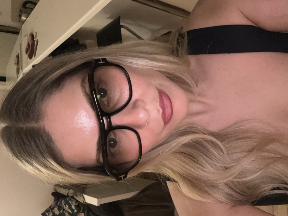 A woman with fair skin, light blonde hair, and wearing large black framed glasses gazes directly at the camera. Her lips are painted a reddish-pink. A black strap is visible near her shoulder. The background includes a shelf and other indistinct objects. The image is rotated, placing the woman's head in the top left corner.