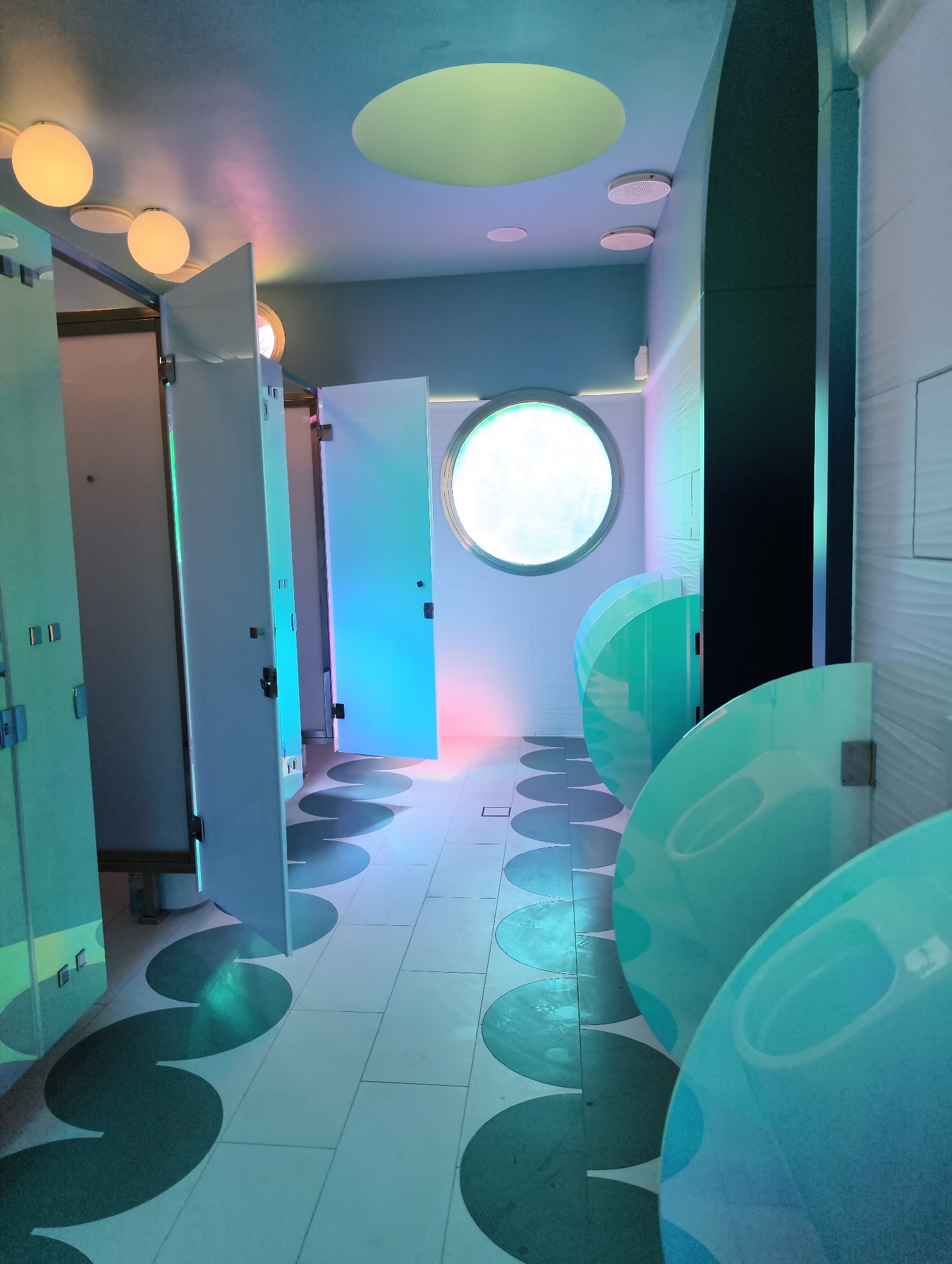 This image captures a wide-angle view of a distinctly modern and brightly lit public restroom in Krasnodar, Russia. The scene is empty of people, suggesting either a time outside of peak use or a recently cleaned facility.
The restroom features several private cubicles with white doors, some slightly ajar, visible on the left side. Beyond these, a row of translucent, light blue-green urinals with rounded dividers extends along the right wall. The floor is tiled, predominantly white, with a striking pattern of large, dark circular shapes that give a playful or artistic feel to the space.
A prominent feature is the lighting, which casts a vibrant, multi-colored glow across the room. Overhead, several spherical ceiling lights emit a warm, orange-yellow light, contrasting with cool blue and pink washes that illuminate the walls and reflect on the floor and fixtures. A large, circular window, glowing brightly with an unseen light source, is centered on the back wall, resembling a porthole. Other ceiling elements include recessed lights and white circular vents. The walls vary, with some being smooth and plain, while others, particularly behind the urinals, exhibit a subtle wave-like texture. A tall, dark column or wall section stands vertically on the right, providing a strong linear contrast to the numerous circular elements.
No specific activity is taking place, and the restroom appears to be in pristine condition. There is no visible text in the image. The overall impression is clean, contemporary, and somewhat futuristic, utilizing a blend of geometric shapes and dynamic lighting.
