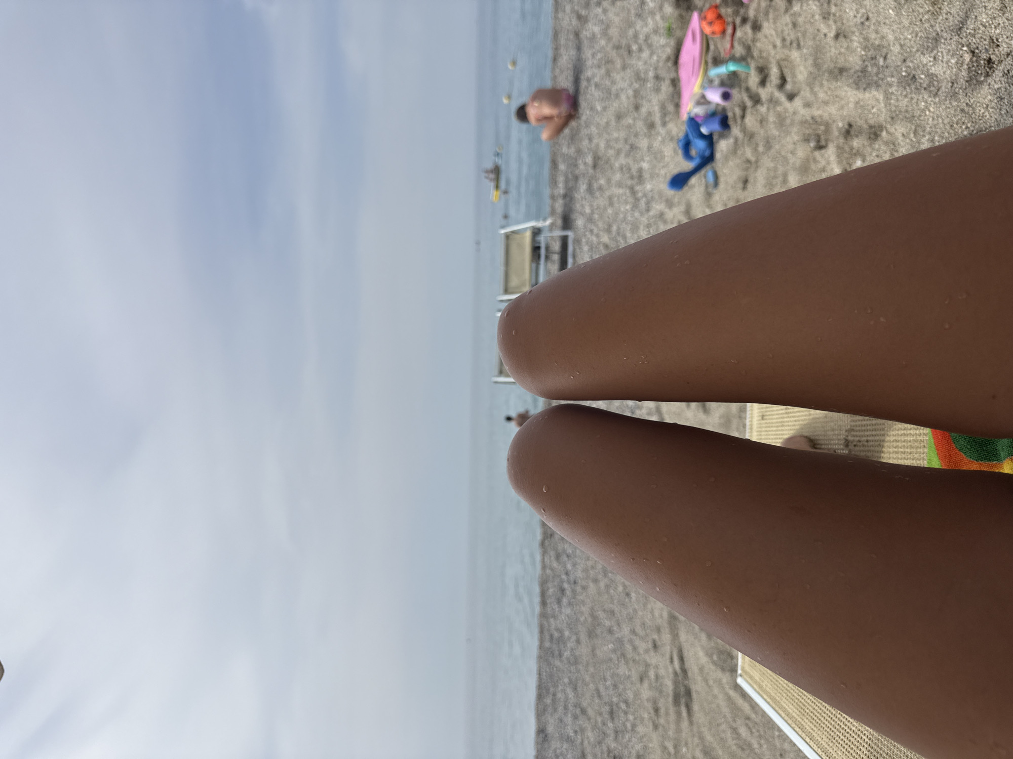 This is a first-person perspective image taken at a beach, likely in Finale Ligure, Italy. The foreground is dominated by the legs of a person lying on a beach chair, with small water droplets visible on their skin. The beach is covered in pebbles, and beyond it, the calm blue sea stretches to the horizon under a cloudy sky. In the middle ground, several beach chairs are set up, and two individuals are visible. One person is partially submerged in the water, and another is sitting on the beach. In the background, a few small boats are dotted on the water, and the sky is overcast. Scattered on the sand in the foreground are several beach toys, including a pink swim fin, blue inflatable toys, and a small orange ball. The overall scene depicts a relaxed day at the beach.