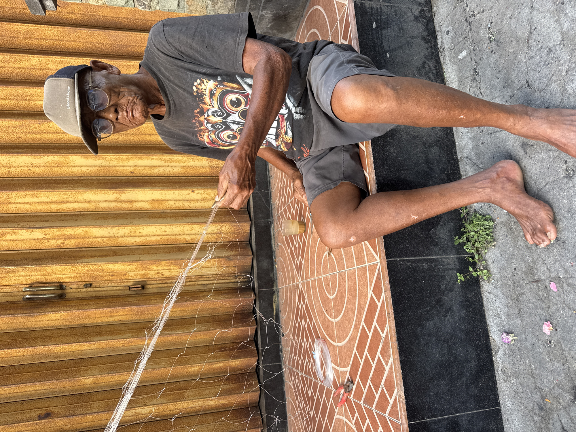 An elderly Indonesian man, wearing a dark t-shirt, grey shorts, glasses, and a baseball cap, is sitting on the tiled step of a building. He is working on a white fishing net, holding the strands in his hands. Next to him on the step are a pair of red-handled pliers and a spool of white line. A small clear cup containing a light-colored liquid is also on the step. The background features a closed, corrugated metal door with vertical lines. The ground in front of the step is paved, and a few fallen flower petals are visible. The scene appears to be in a street or outdoor area in Kuta, Indonesia, suggested by the style of the building and the presence of the fishing net. The man's skin is weathered, and he has some visible scars on his legs. The lighting indicates it is daytime, with sunlight casting shadows.