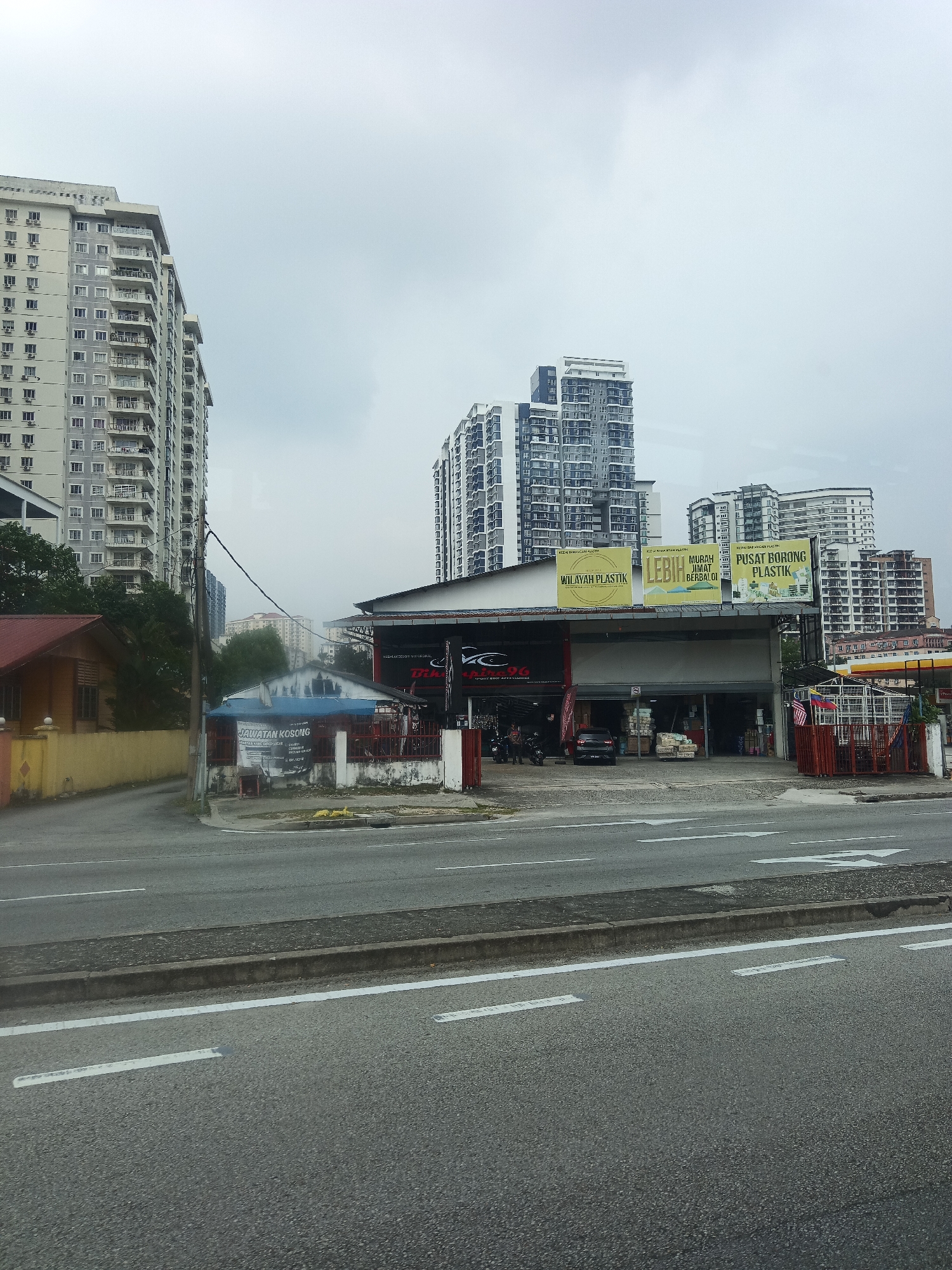 The image captures a commercial building with a bright yellow awning, located in Kuala Lumpur, Malaysia. The building appears to be a retail establishment, possibly a warehouse or a wholesale store, as indicated by the signs displaying "PUSAT BORONG PLASTIK" (Plastic Wholesale Center) and "LEBIH MURAH JIMENT BERBALOI" (Cheaper, More Economical, Worthwhile). These signs are in Malay. Several vehicles, including a black car and motorcycles, are parked or present near the entrance of the building. The establishment is situated alongside a multi-lane road with clear white lane markings, suggesting it is in a busy urban area. In the background, multiple high-rise buildings and apartment complexes dominate the skyline, characteristic of Kuala Lumpur's urban landscape. The sky is overcast, contributing to a diffused lighting condition. A smaller, single-story house with a red roof is visible to the left of the commercial building. The scene suggests a typical commercial street in the Malaysian capital.