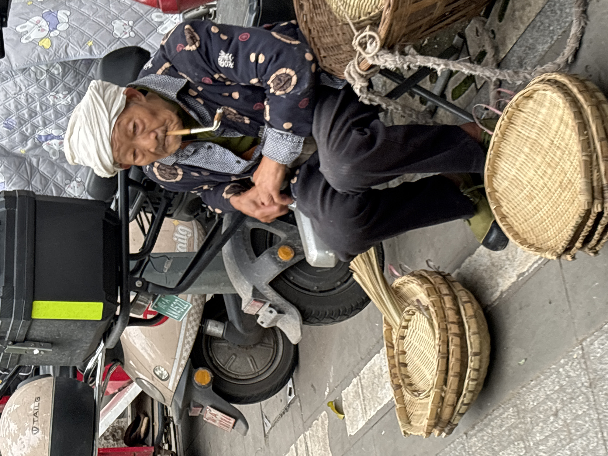 An elderly woman with a white headscarf is sitting on an electric moped on a city street in Jiangjin, China. She is wearing a dark patterned jacket and dark pants. A smoking pipe is held in her mouth. The moped is beige with a black storage box attached. Baskets are stacked around the moped. A license plate on the moped reads "AN45222". The ground is paved with gray stones.