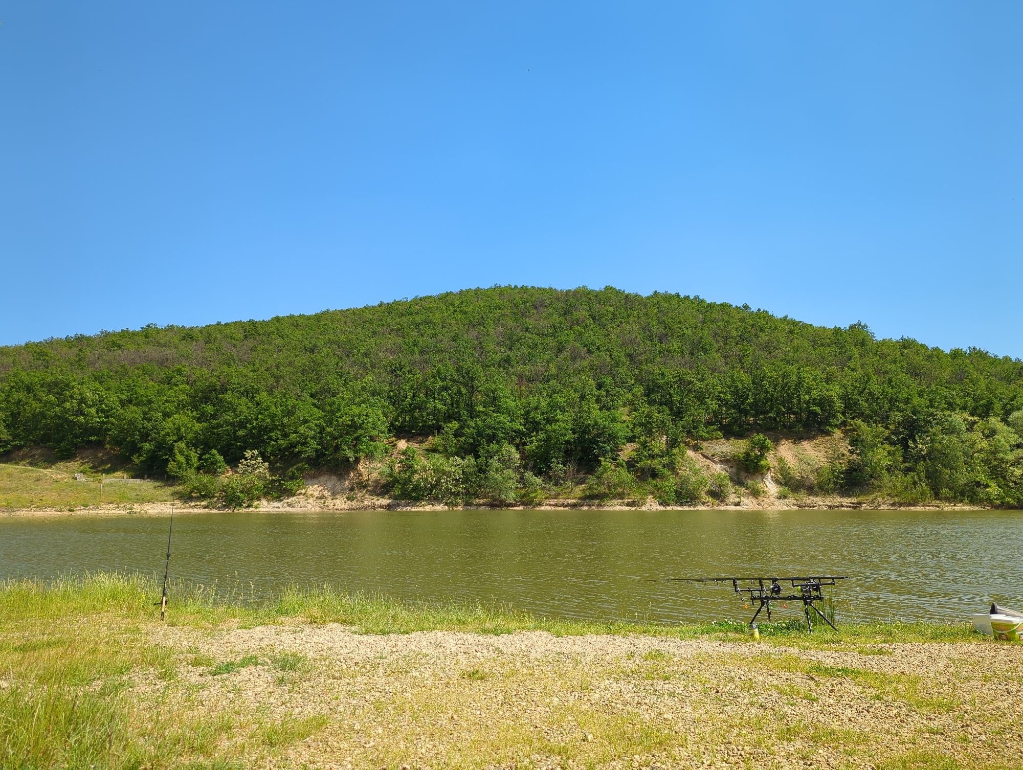 The image depicts a fishing scene near Slavovitsa, Bulgaria. A body of water fills the midground, with a tree-covered hill rising in the background under a clear blue sky. On the grassy and gravelly shoreline, fishing equipment is set up: a rod stands to the left, and a rod pod supporting multiple rods stands near the center-right, with a container to the far right.