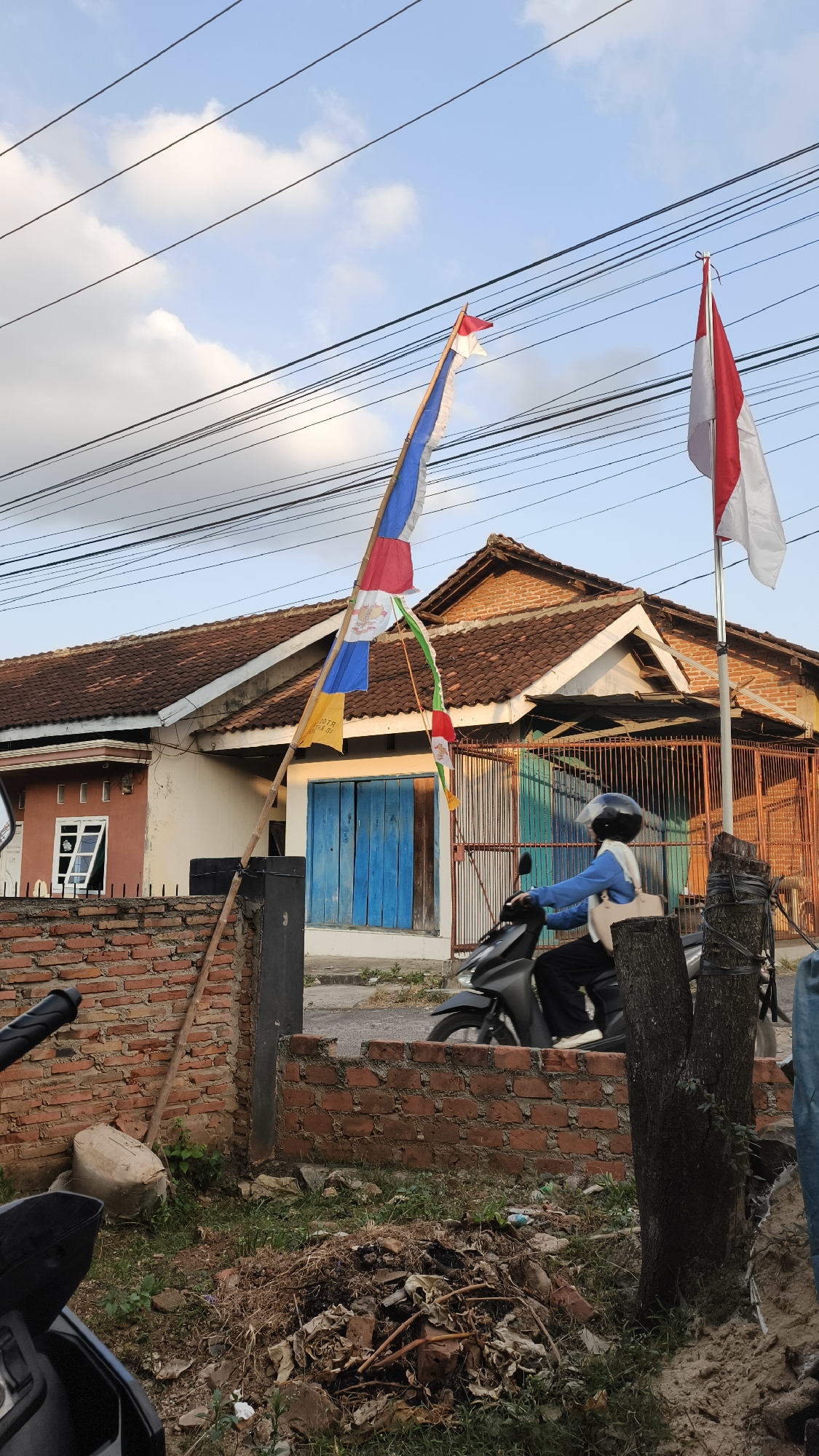 The image depicts a street scene in Bandar Lampung, Indonesia, likely during daylight hours given the lighting conditions and visible shadows. In the foreground, a motorcycle with a rider is partially visible on the right side of the frame, suggesting movement or stationary presence on a paved surface. A pile of dry vegetation and debris is present on the ground in the mid-ground, adjacent to a short, red brick wall. A large tree trunk stands to the right of the debris pile.
Behind the wall, a house with a reddish-brown tiled roof is visible. The house has an orange facade on the left and a section with a beige facade on the right. A window with white-framed panes is open on the orange section. A set of bright blue wooden doors is prominent on the beige section. A rusty orange metal fence runs along the front of the property.
Two flags are displayed prominently on a wooden pole erected near the front of the property. The pole is angled and appears to be made of bamboo. The flag on the right is the Indonesian national flag (Merah Putih), red on top and white on the bottom. The flag on the left is a multi-colored banner with red, blue, white, and green sections, and appears to have an emblem or image on it. Another small yellow flag with text is also visible hanging from the pole. Overhead, a network of electrical wires is strung across the sky. The sky is blue with scattered white clouds. The overall scene suggests a residential area in Bandar Lampung, possibly decorated for a national holiday or celebration.